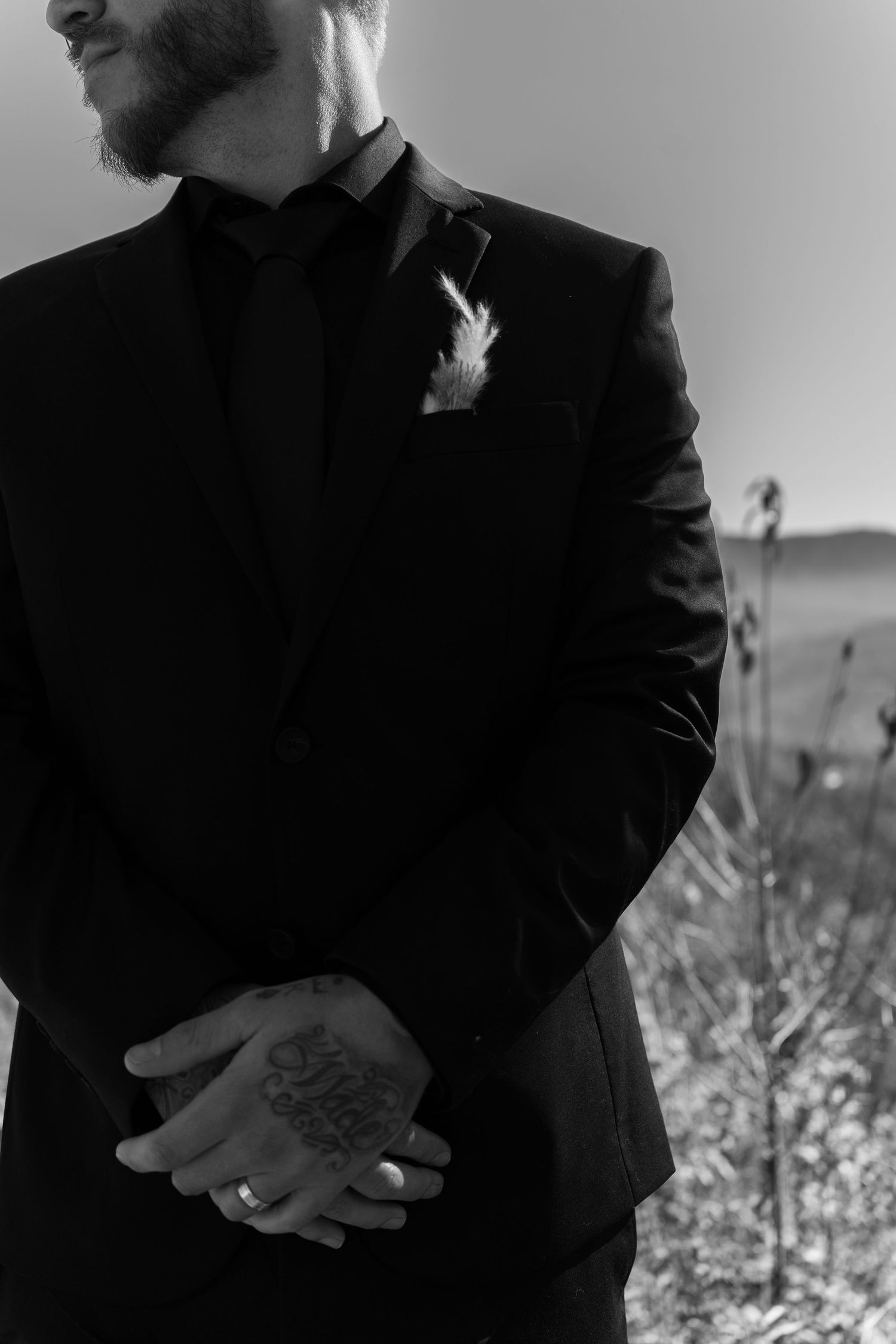 Black and white photo of a groom in a dark suit with boutonniere, hands clasped showing tattoos and wedding ring.