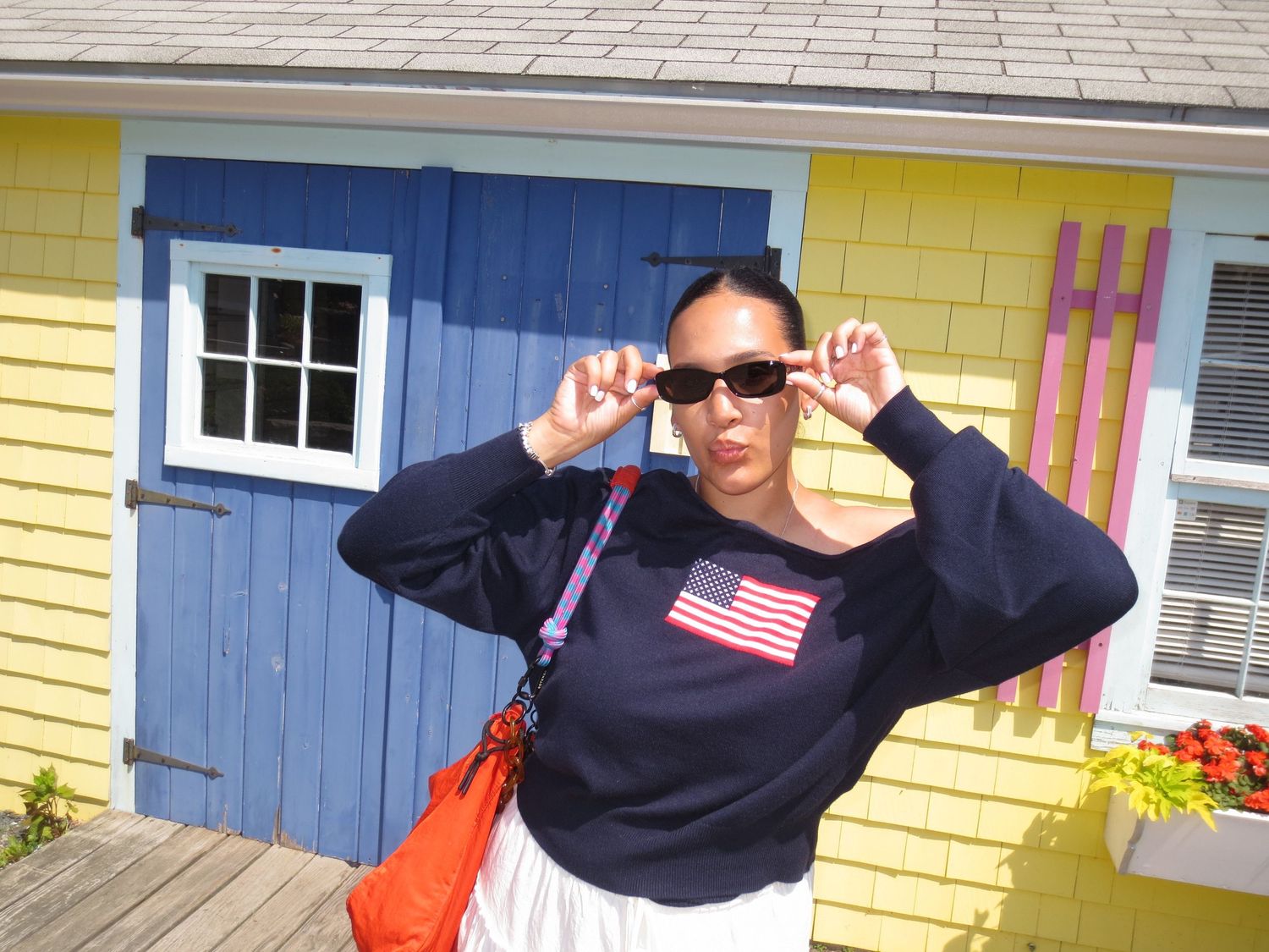 Woman in American flag sweatshirt and sunglasses posing in front of colorful yellow and blue beach house.