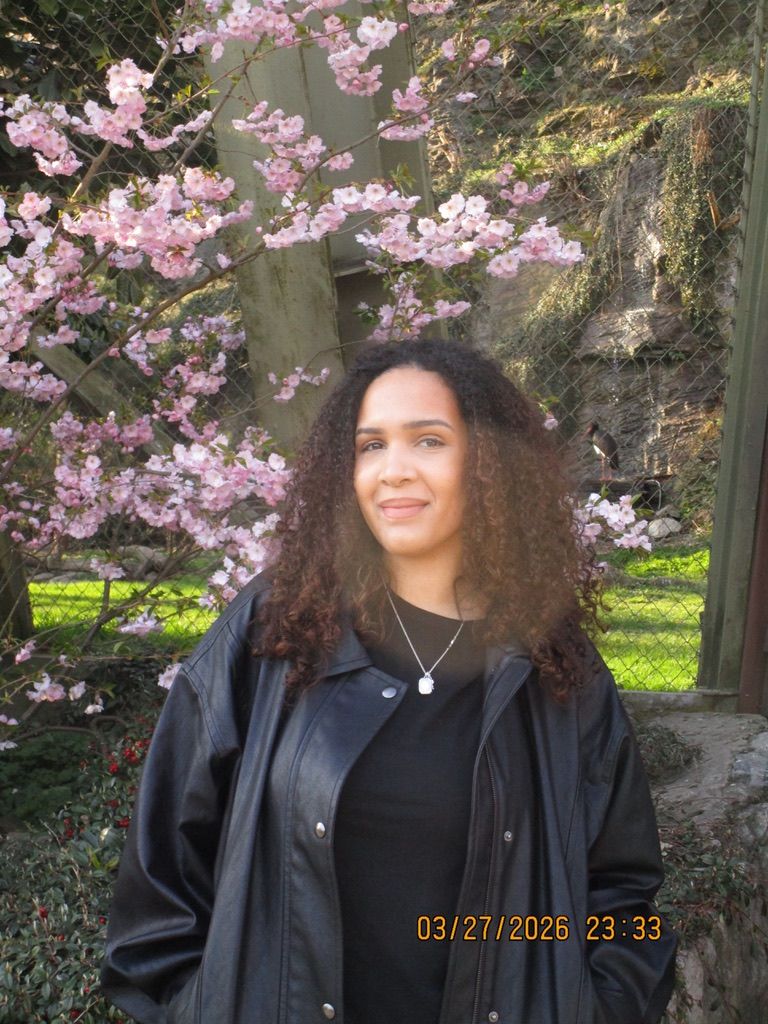 Young woman with curly hair in black leather jacket poses before blooming cherry blossom tree in spring garden.
