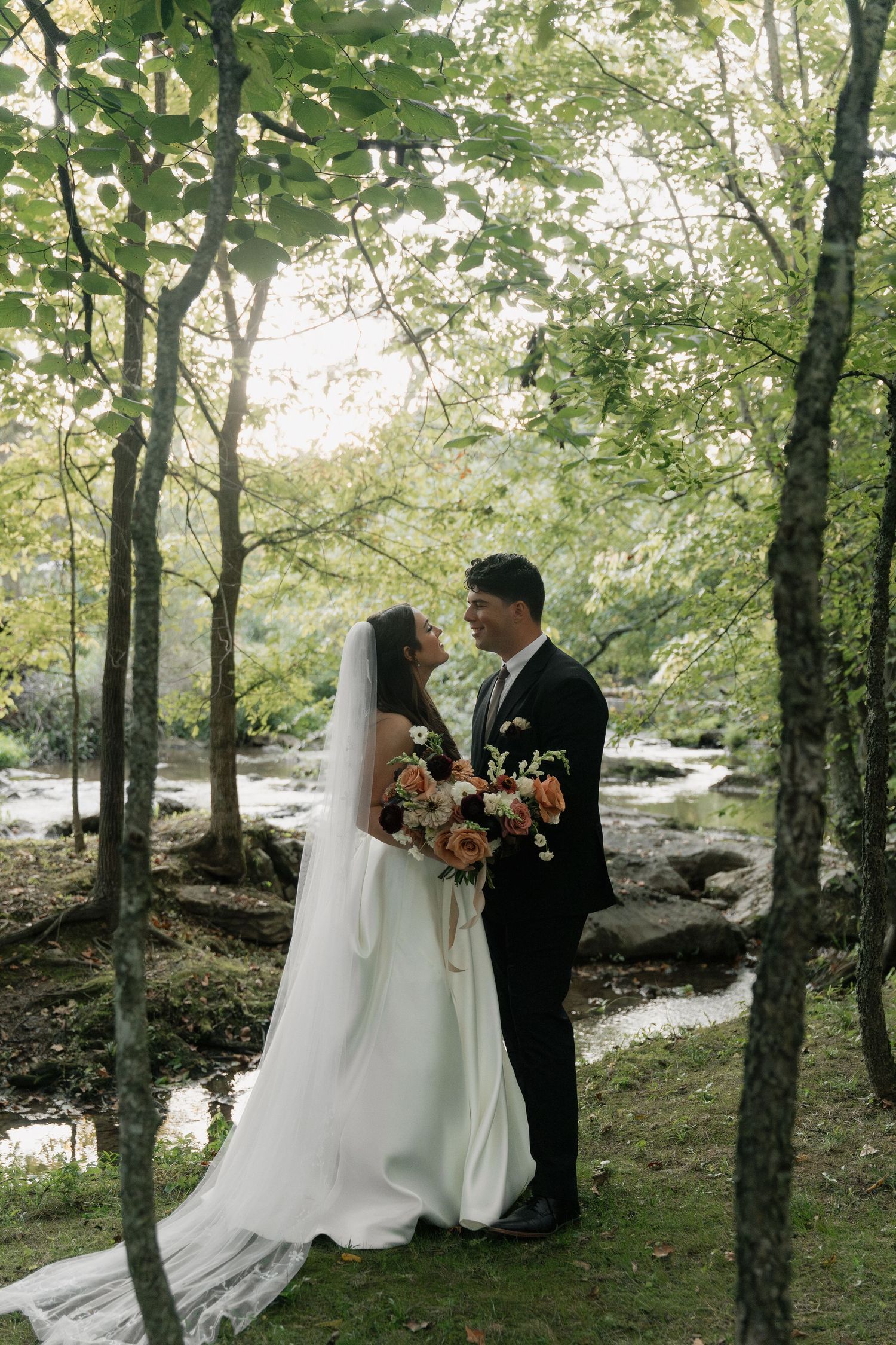 Bride and groom stand among lush green trees near a rocky stream, bride holding a bouquet of peach and orange flowers.