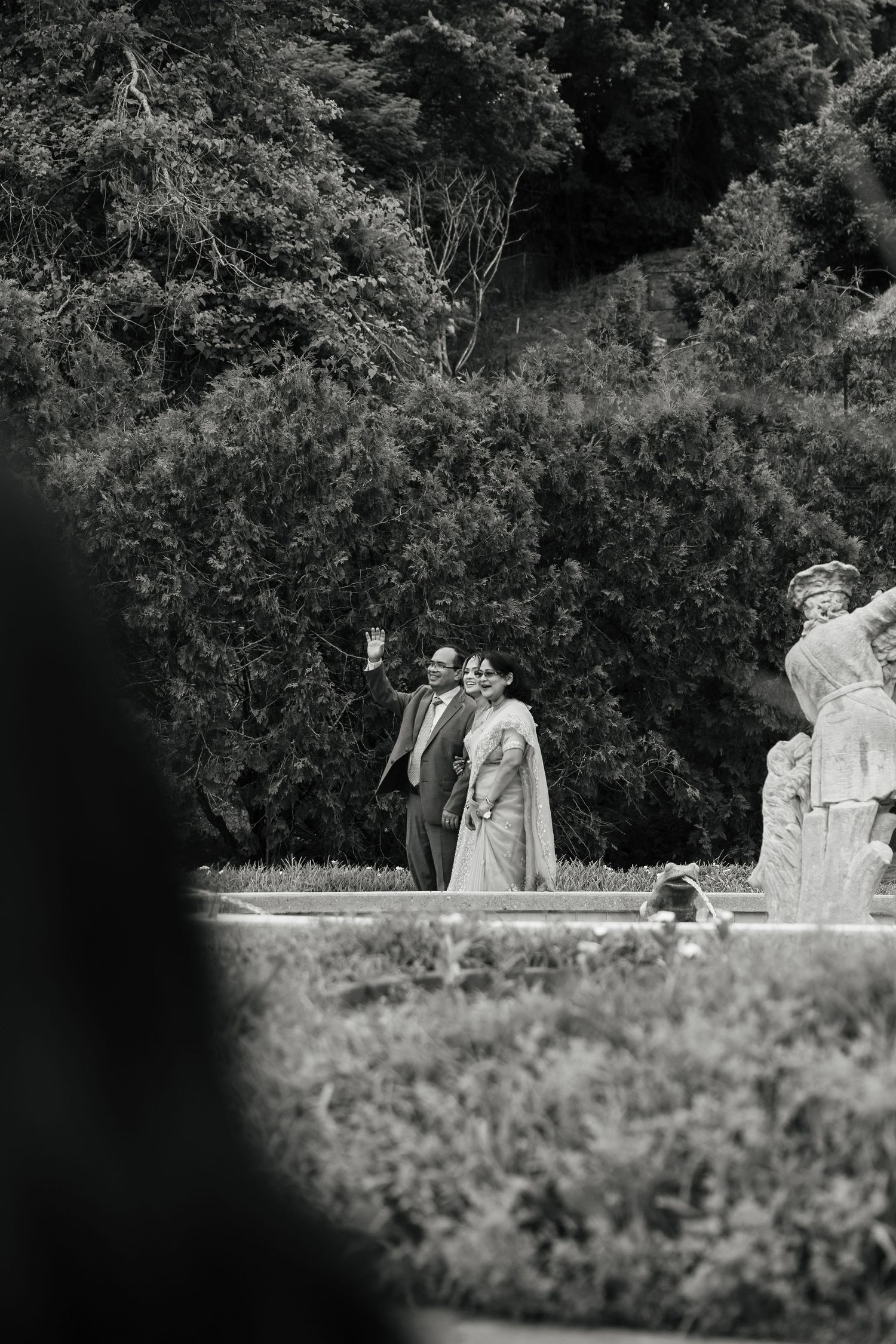 Black and white photo of a couple in formal attire waving in a garden with lush trees and statues.