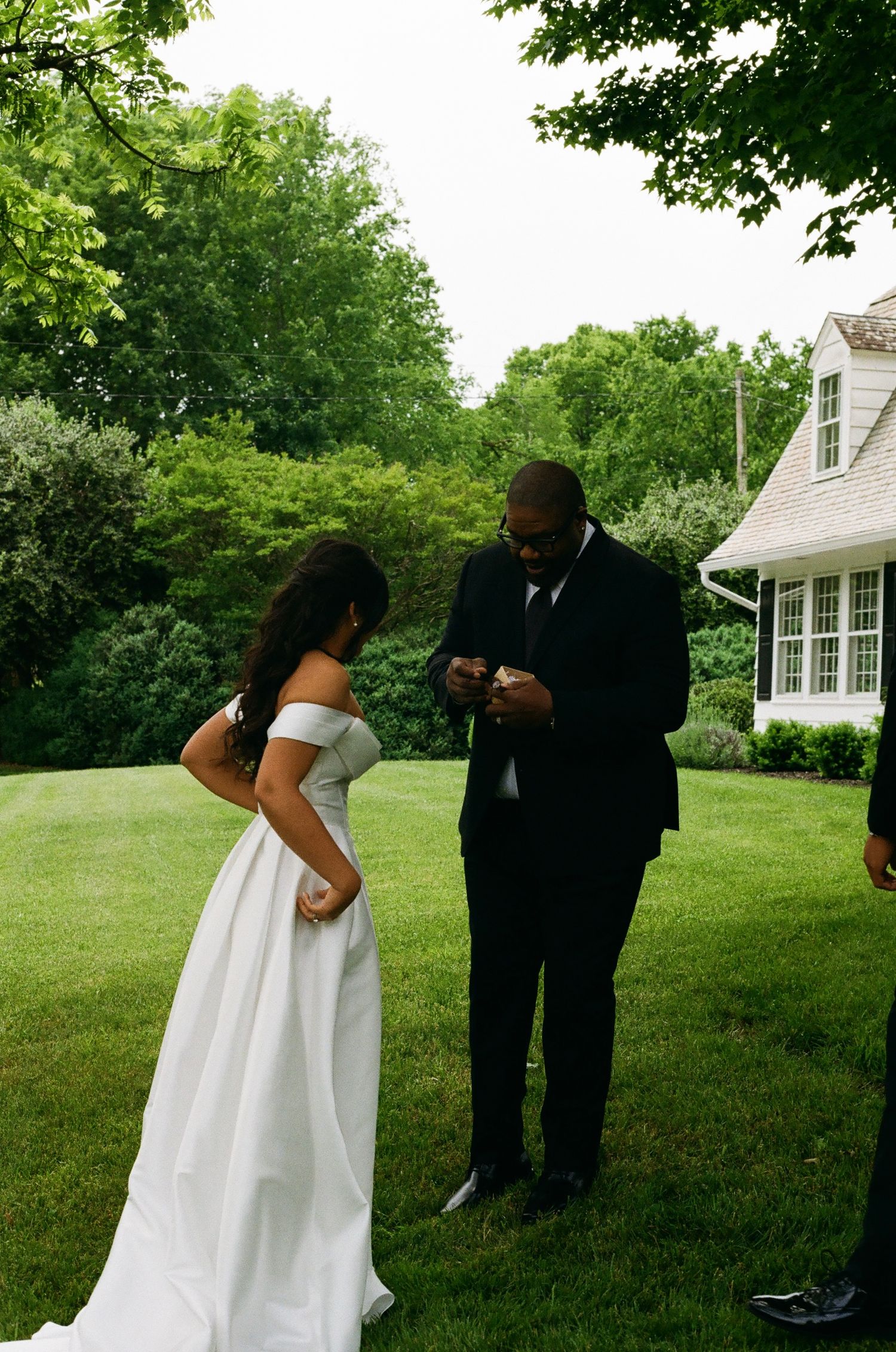 Bride in white off-shoulder gown and groom in black suit exchange rings outdoors on green lawn near white house.