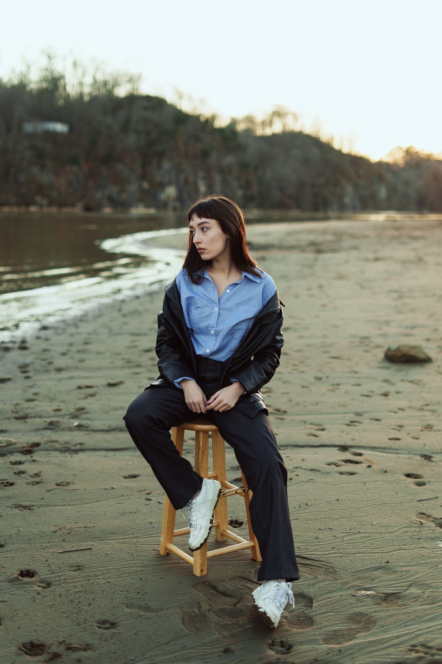 Someone sitting on a wooden stool at the beach wearing a blue button-down shirt and black pants at sunset.