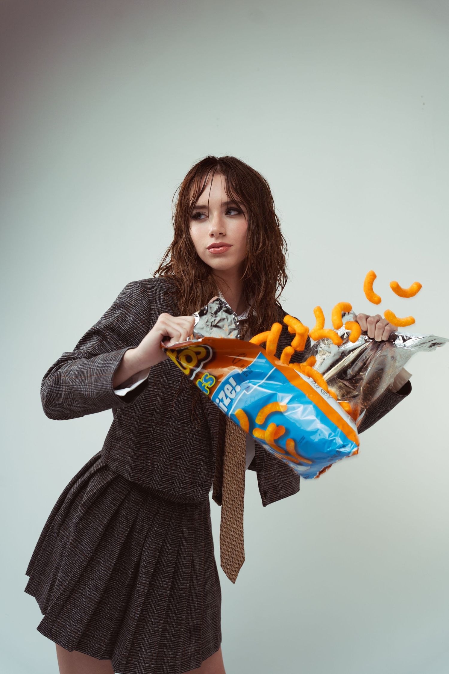 A figure in a dark dress and blazer poses with a bag of snacks spilling orange cheese puffs against a gray backdrop.
