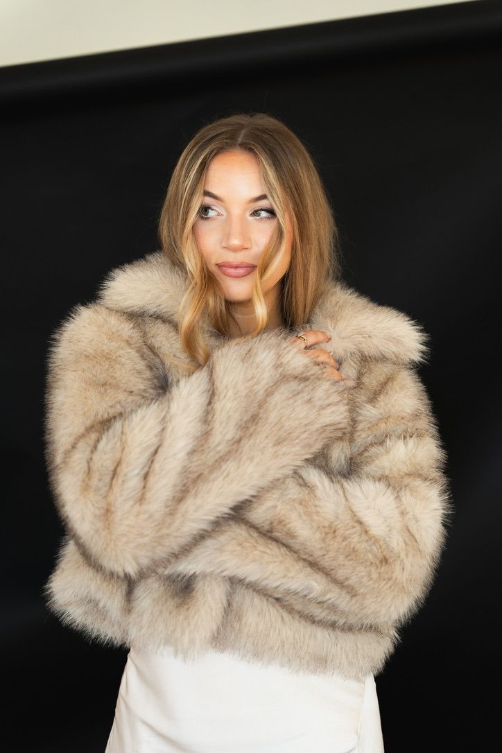 Young woman wearing a luxurious beige fur coat, posing confidently against a dark background.