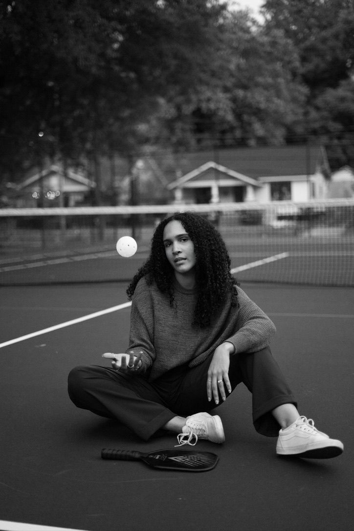 Black and white photo of a young woman with curly hair sitting on a tennis court holding a racket and tossing a ball.