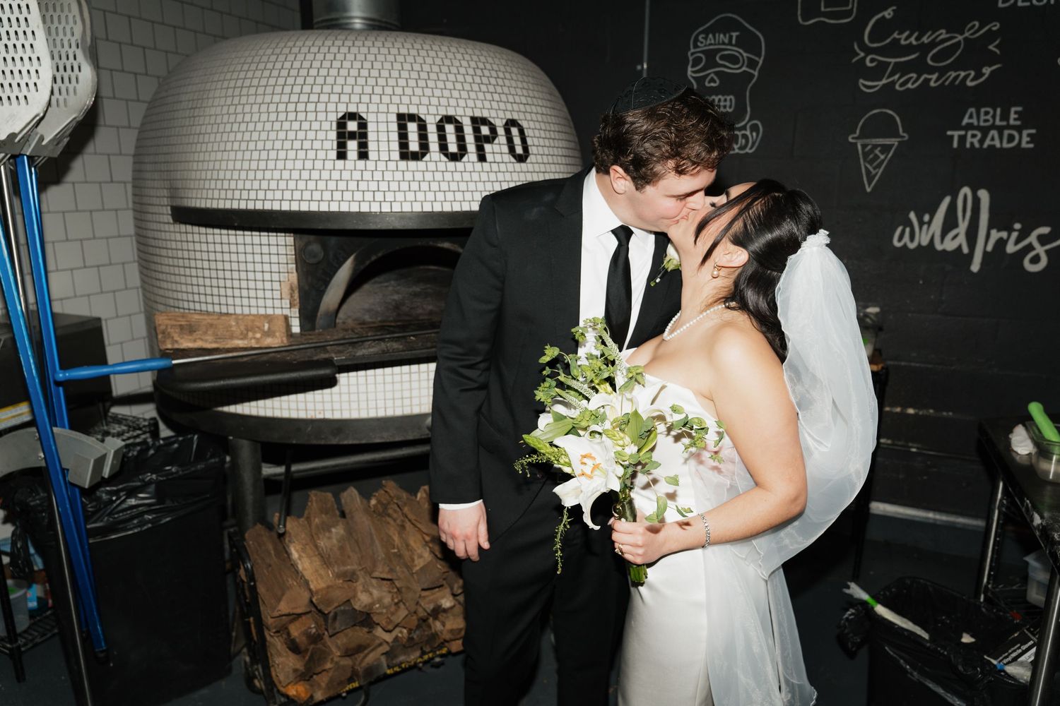 Bride and groom share a kiss in front of a wood-fired pizza oven at A Dopo restaurant wedding venue.