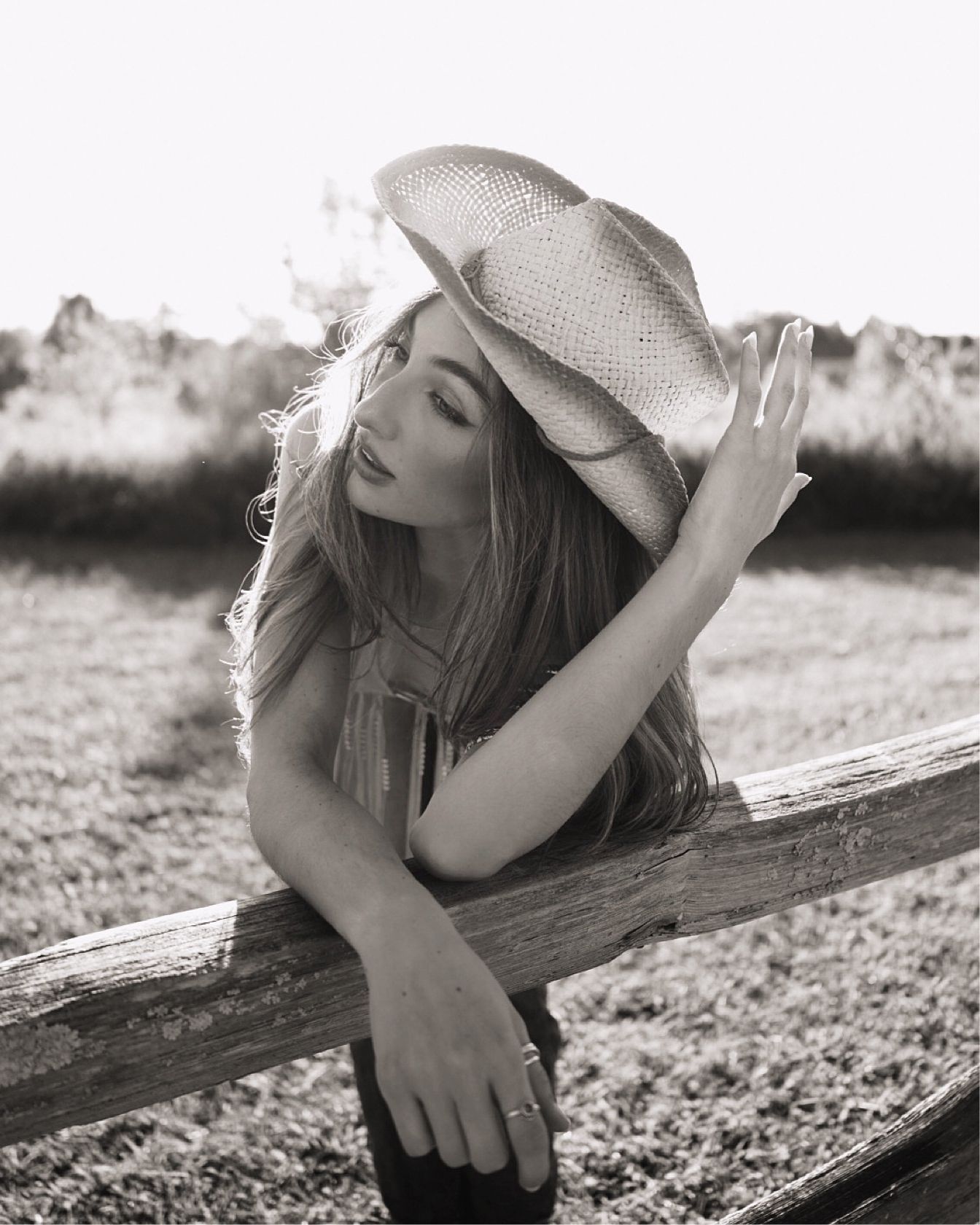 Black and white artistic portrait of someone reclining against a rustic wooden fence wearing a cowboy hat in sunlight.