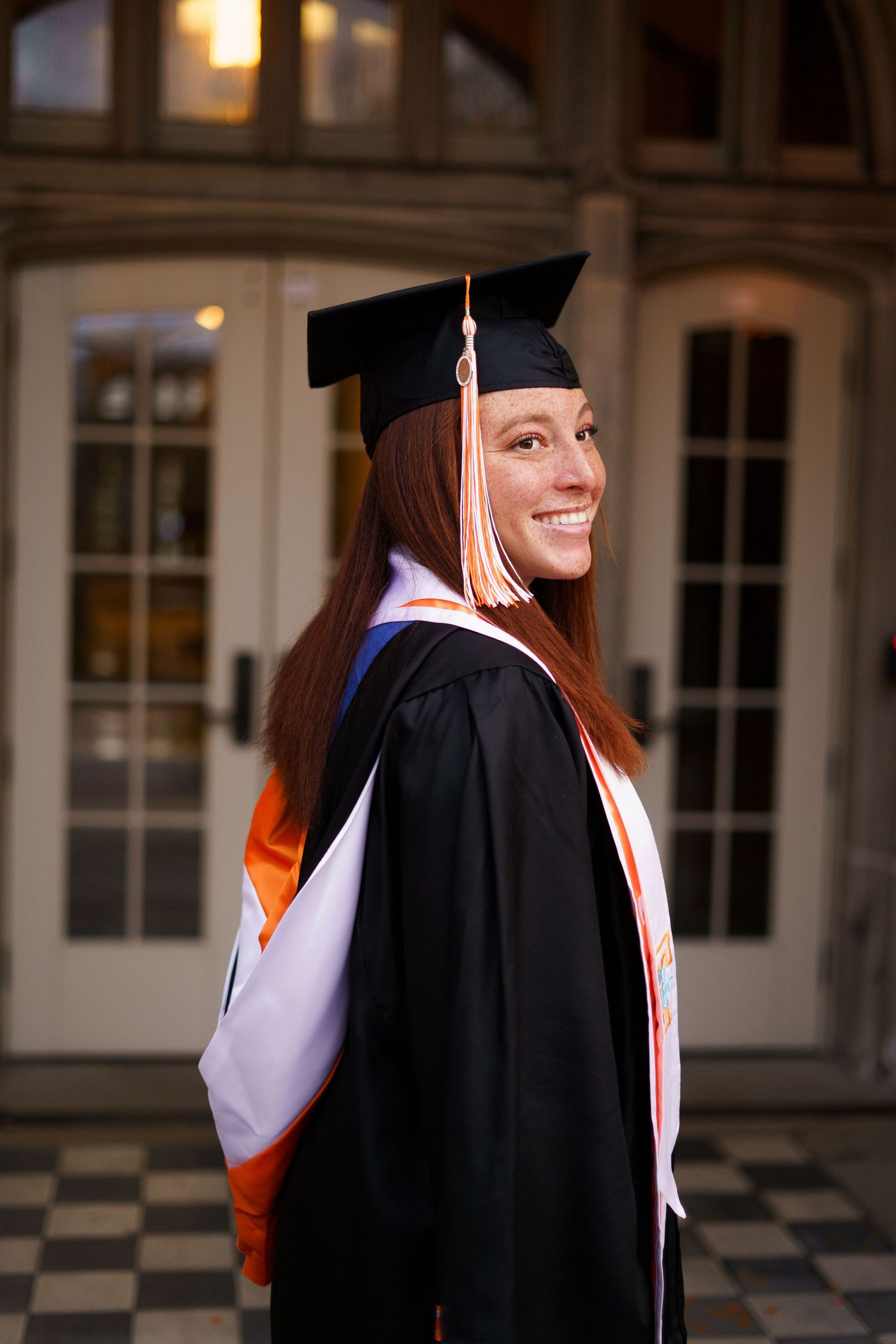 Smiling female graduate in black cap and gown with orange sash poses outdoors.