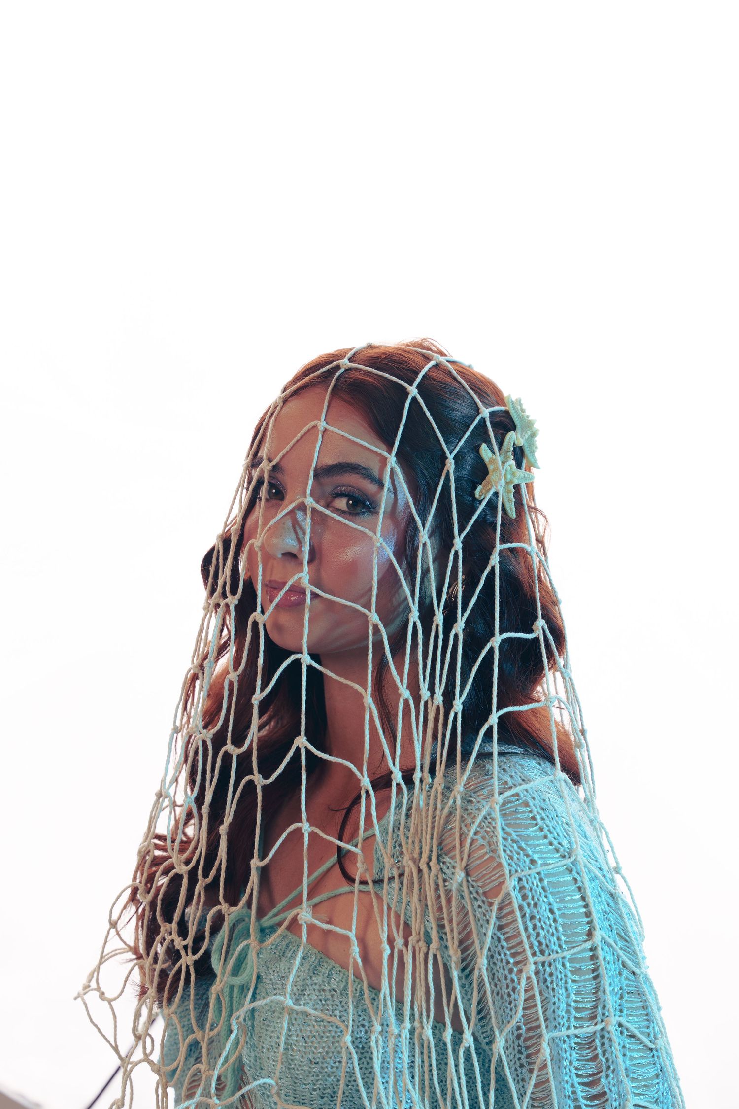 Person wearing turquoise sweater photographed through decorative netting against white background.