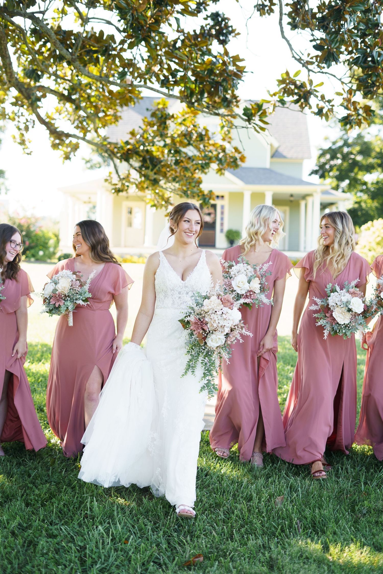 Bride in white lace gown walks with bridesmaids in dusty rose dresses carrying floral bouquets on green lawn.