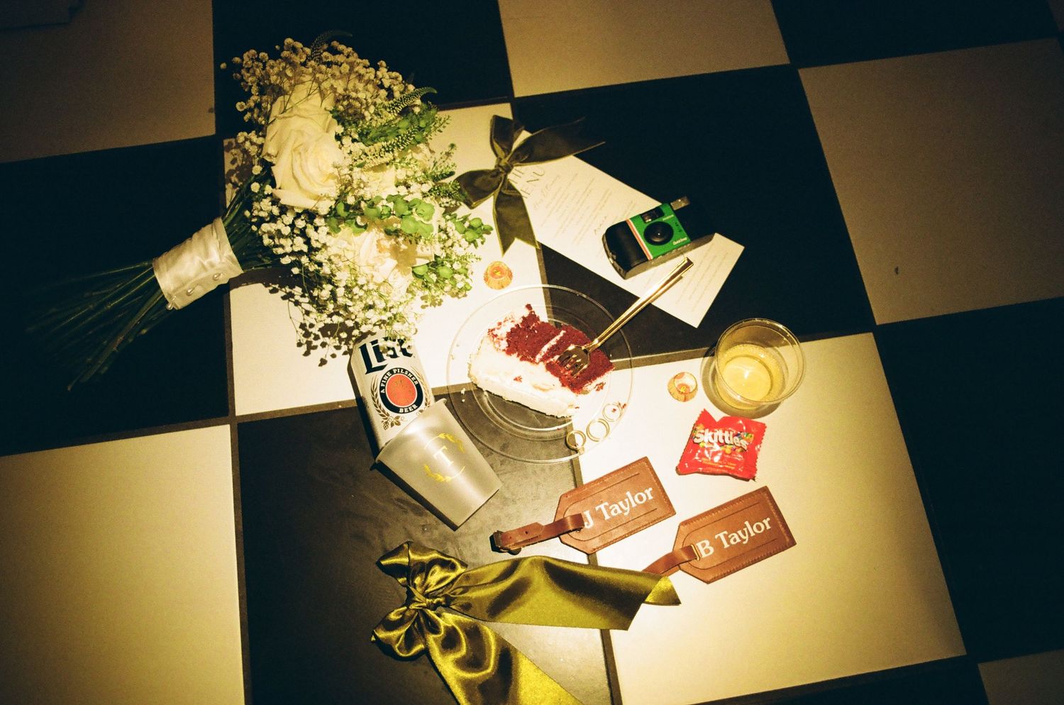 Overhead view of wedding flat lay with white roses, champagne, Taylor gift tags on checkered floor.