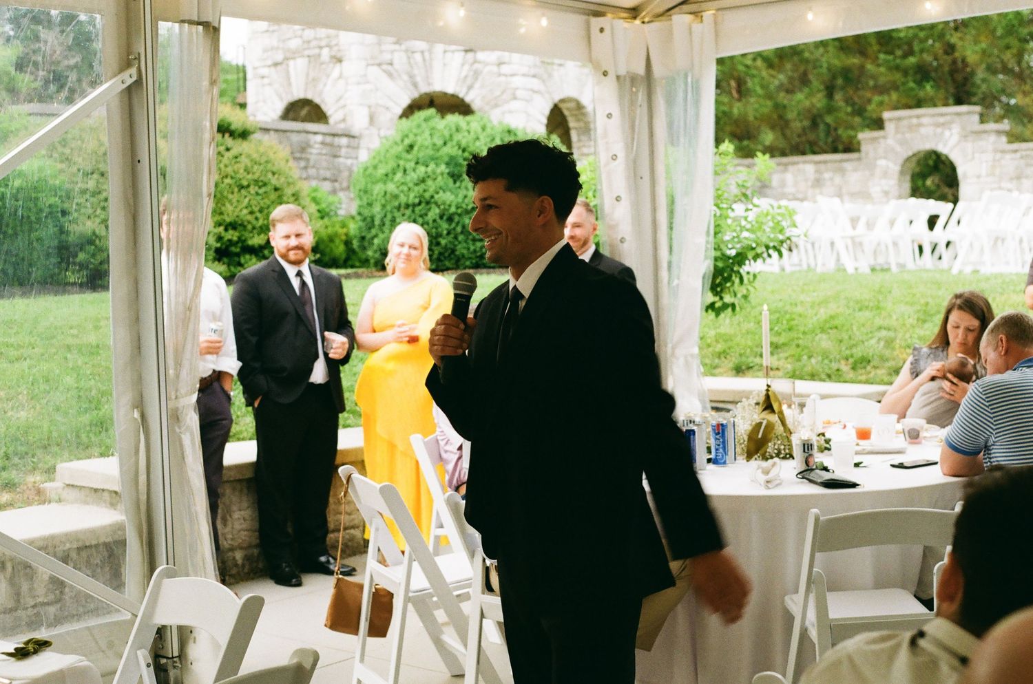 Elegant outdoor wedding reception under a tent with guests in formal attire, including a man in a black suit.