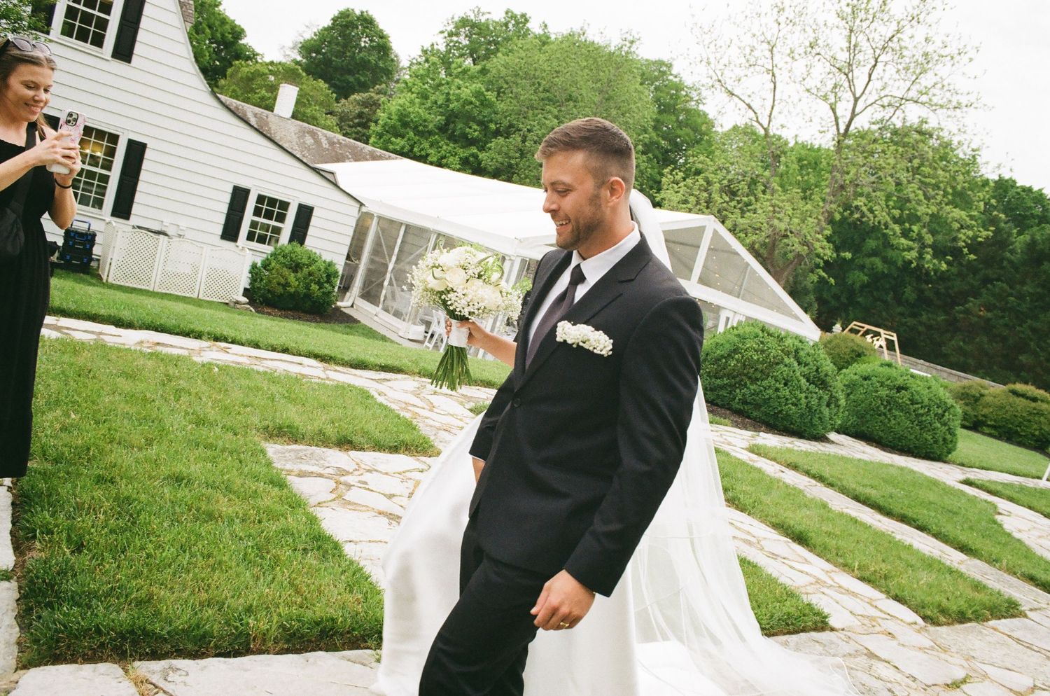 Groom in black tuxedo walks with bride in white gown outside elegant white estate during outdoor wedding ceremony.