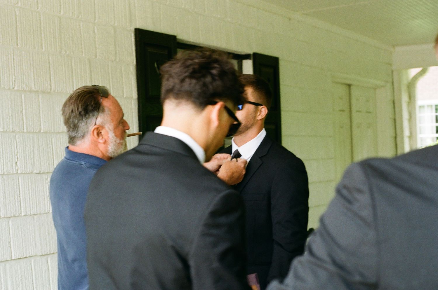 Men in dark suits gathering near a door, with one man in sunglasses having his tie adjusted before a formal event.