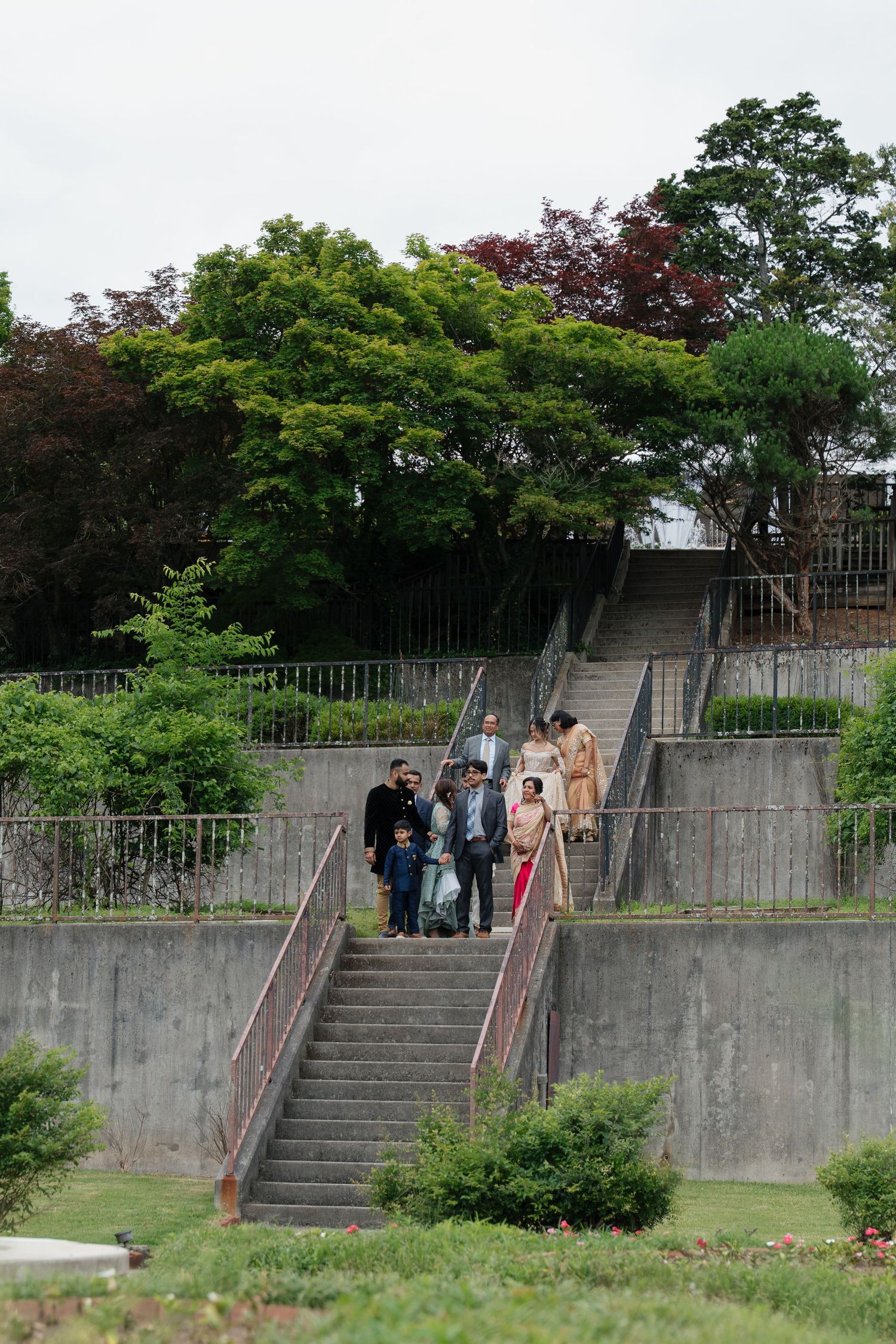 Wedding guests gather on concrete stairs surrounded by lush greenery and trees.