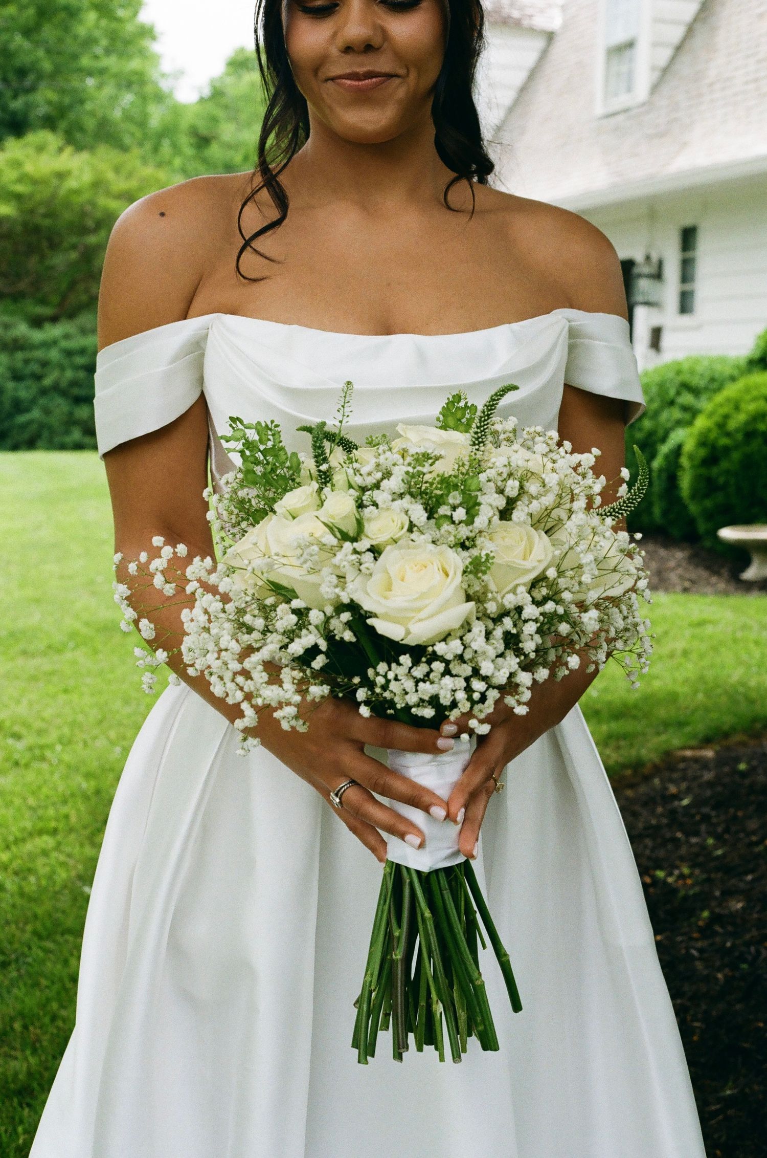 Bride in off-shoulder white gown holding elegant bouquet of white roses and baby's breath outdoors.
