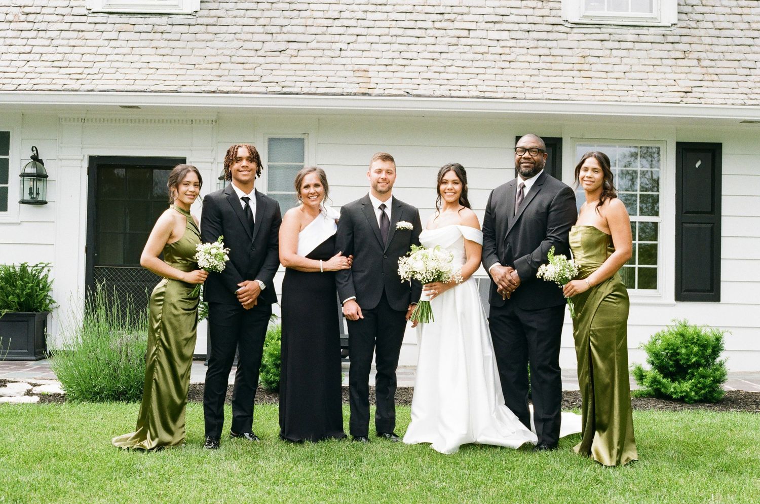 Wedding party posing in front of white house, bride in white gown with groom and bridesmaids in olive green dresses.