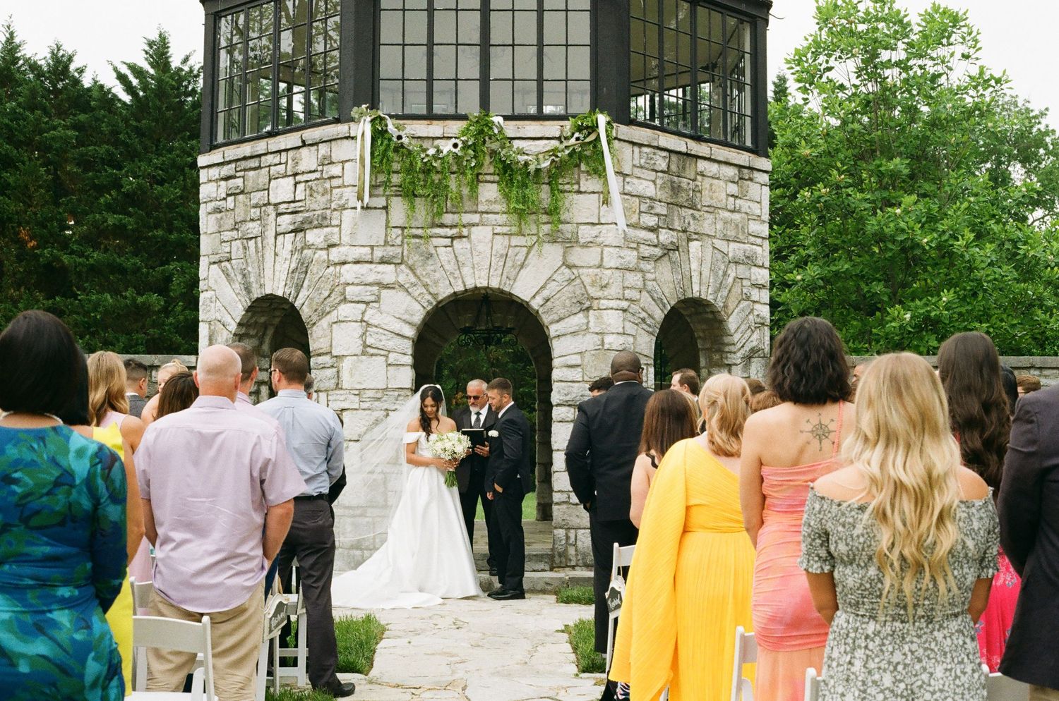 Outdoor wedding ceremony at a stone chapel with guests seated, bride and groom at the altar surrounded by lush greenery.