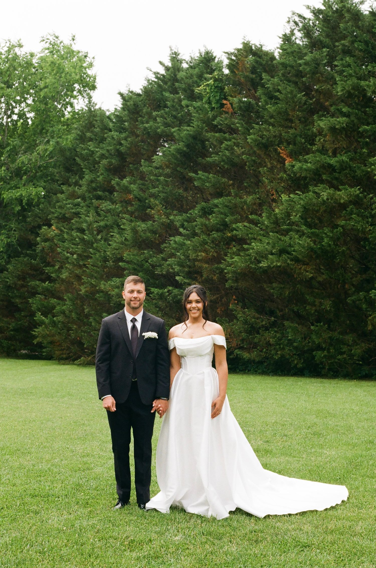 A bride in an off-shoulder white gown and groom in a black suit hold hands on a green lawn with evergreen trees behind them.