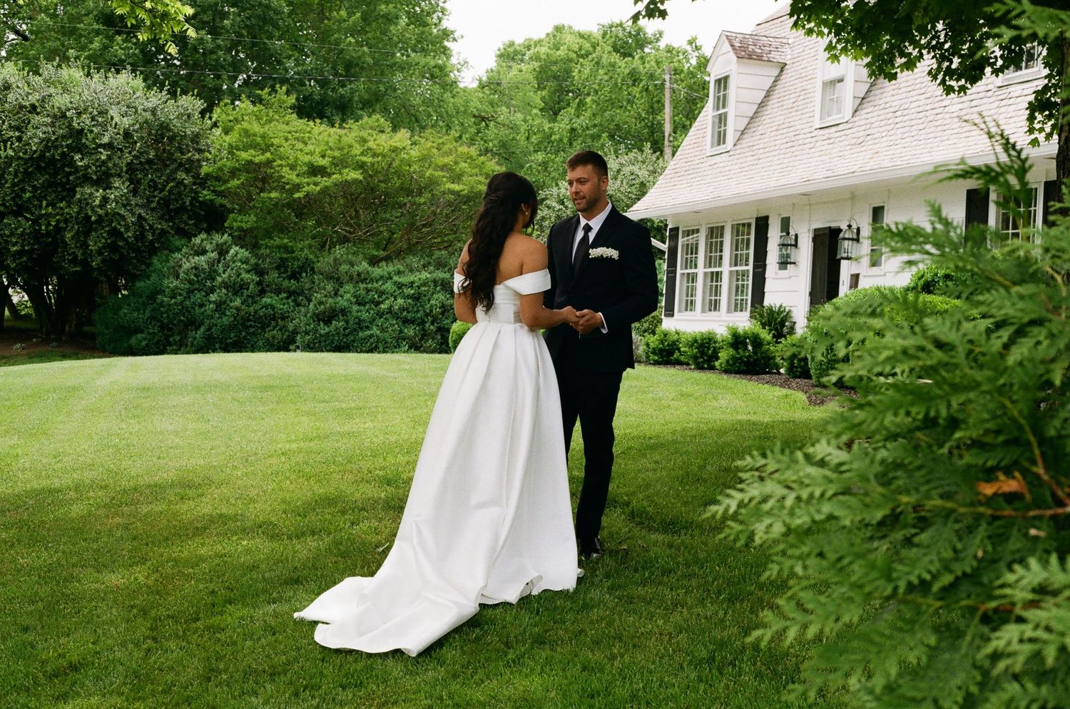 Bride and groom share a romantic first look moment on lush green lawn beside a charming white colonial home.