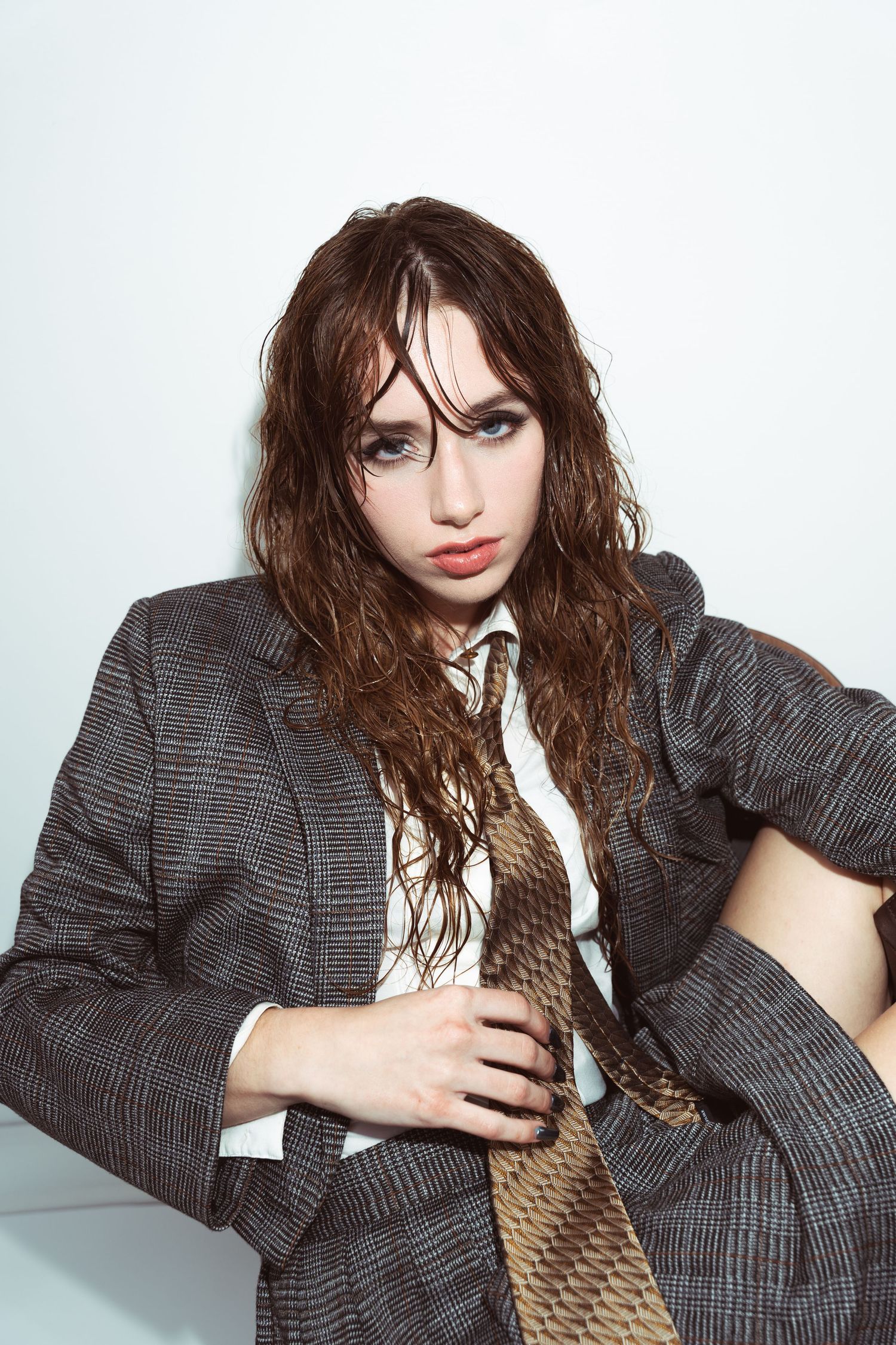 Fashion model wearing oversized plaid blazer with brown tie and tousled wet-look hair against white background.