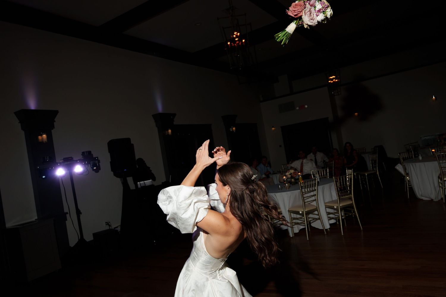 A bride dances alone on the wooden dance floor of a darkened reception hall with stage lights in the background.