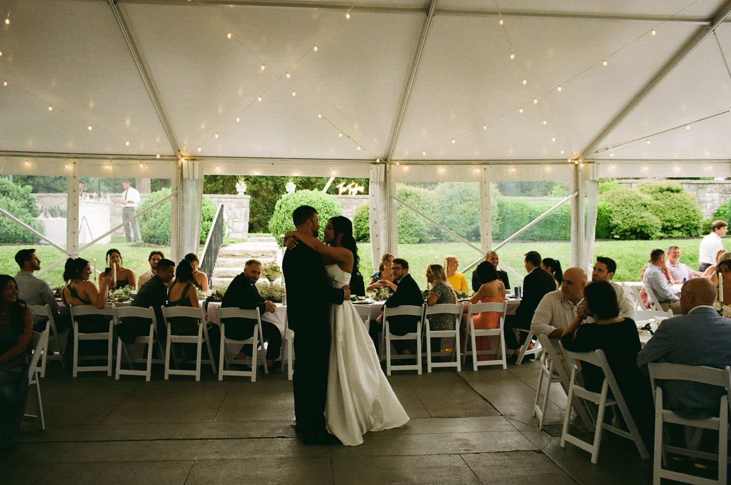 Bride and groom share first dance under elegant tent with string lights at outdoor wedding reception.