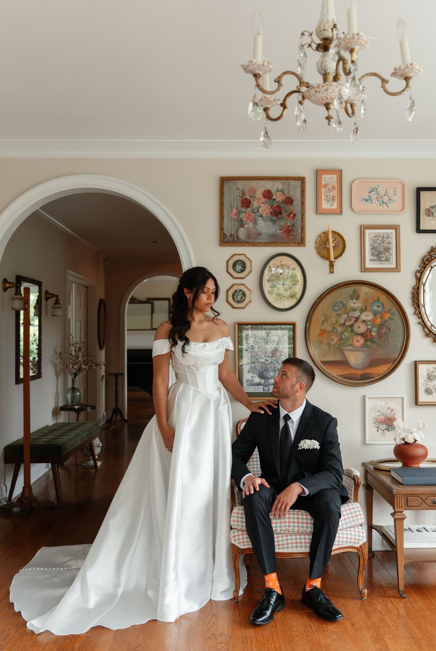Bride in white off-shoulder gown stands beside groom in black suit seated by vintage gallery wall under crystal chandelier.
