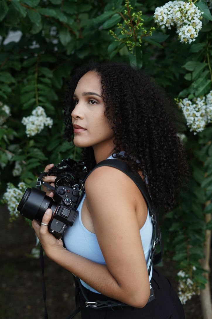 Young woman with curly hair holding a camera outdoors near white flowering bushes.