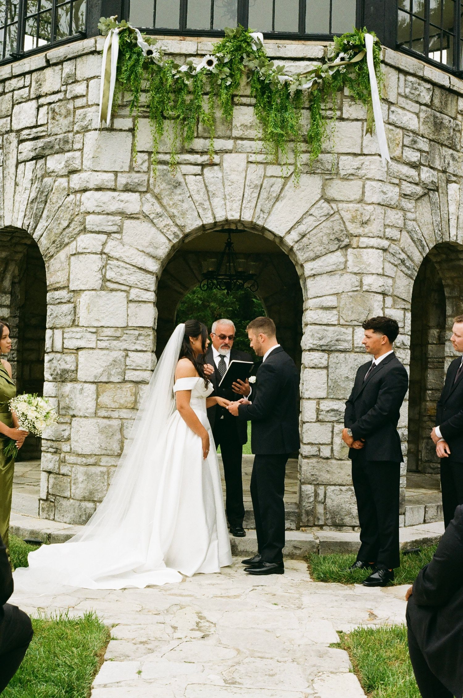 Bride and groom exchange vows outdoors before a stone castle arch adorned with greenery and white flowers.