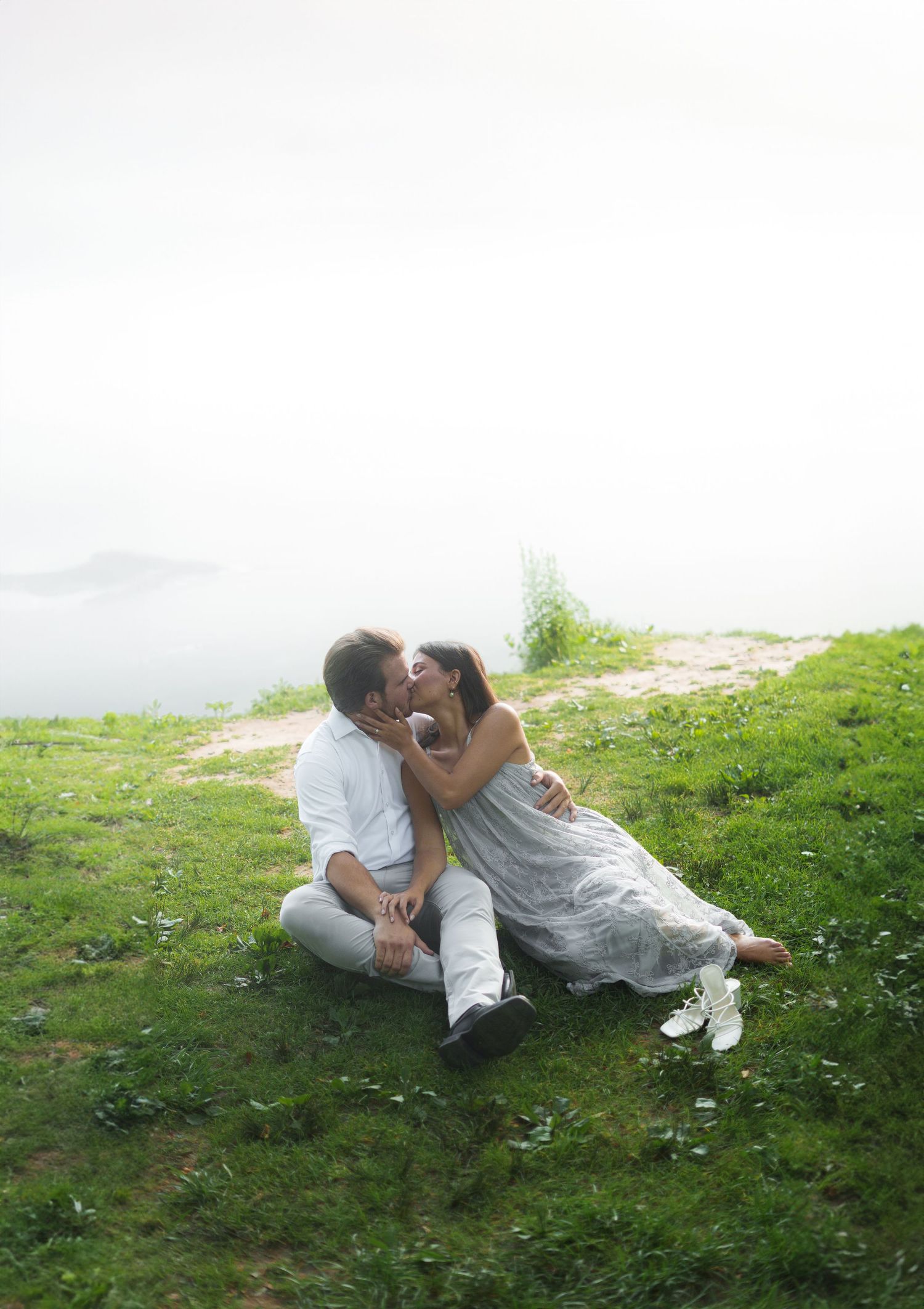Couple sitting on green grass kissing romantically with a misty ocean backdrop on a cloudy day.