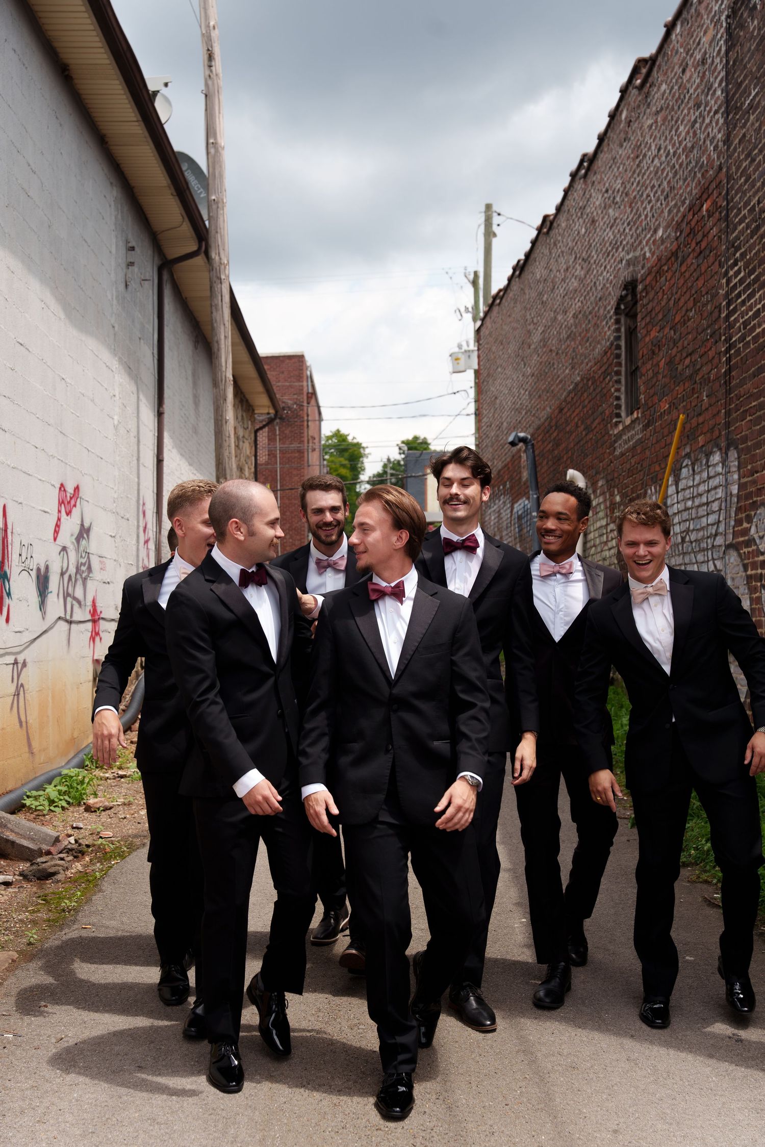 A group in black tuxedos walks together down an urban alleyway in a candid moment.