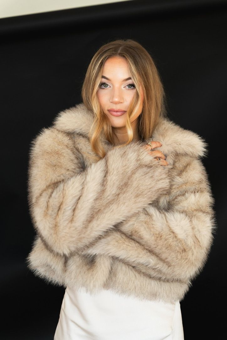 Woman wearing luxurious beige fur coat with arms crossed against dark background.