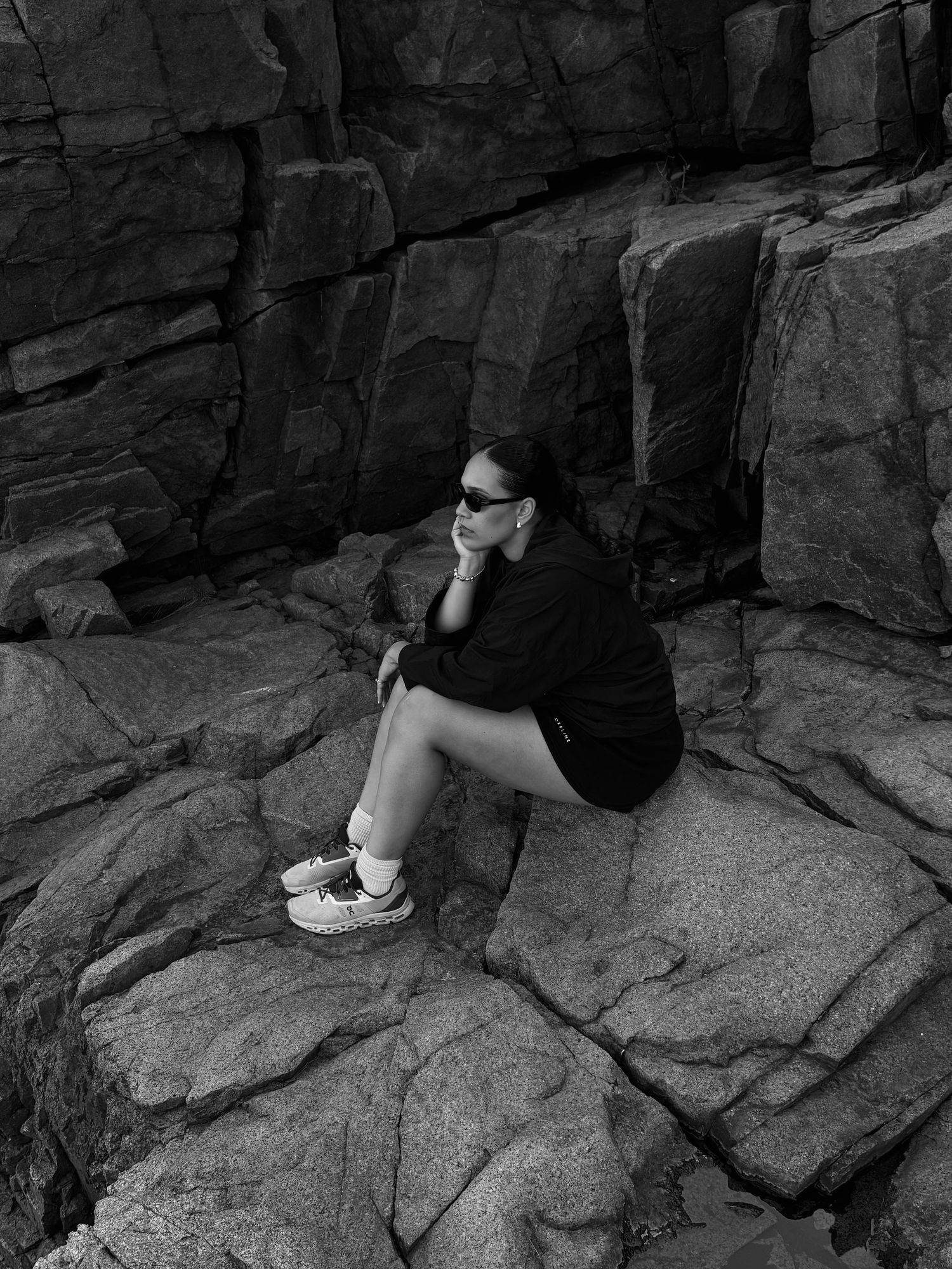 Black and white photo of a woman in sunglasses sitting on rocky terrain, wearing a black outfit and white sneakers.