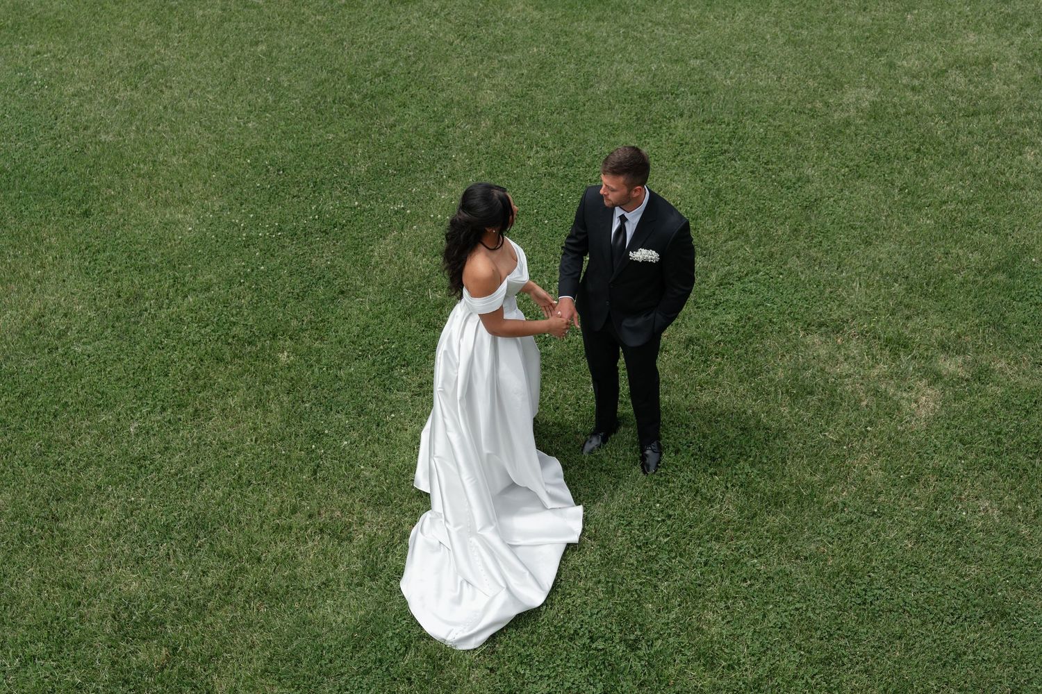 Aerial view of bride in white gown and groom in black suit holding hands on lush green grass.