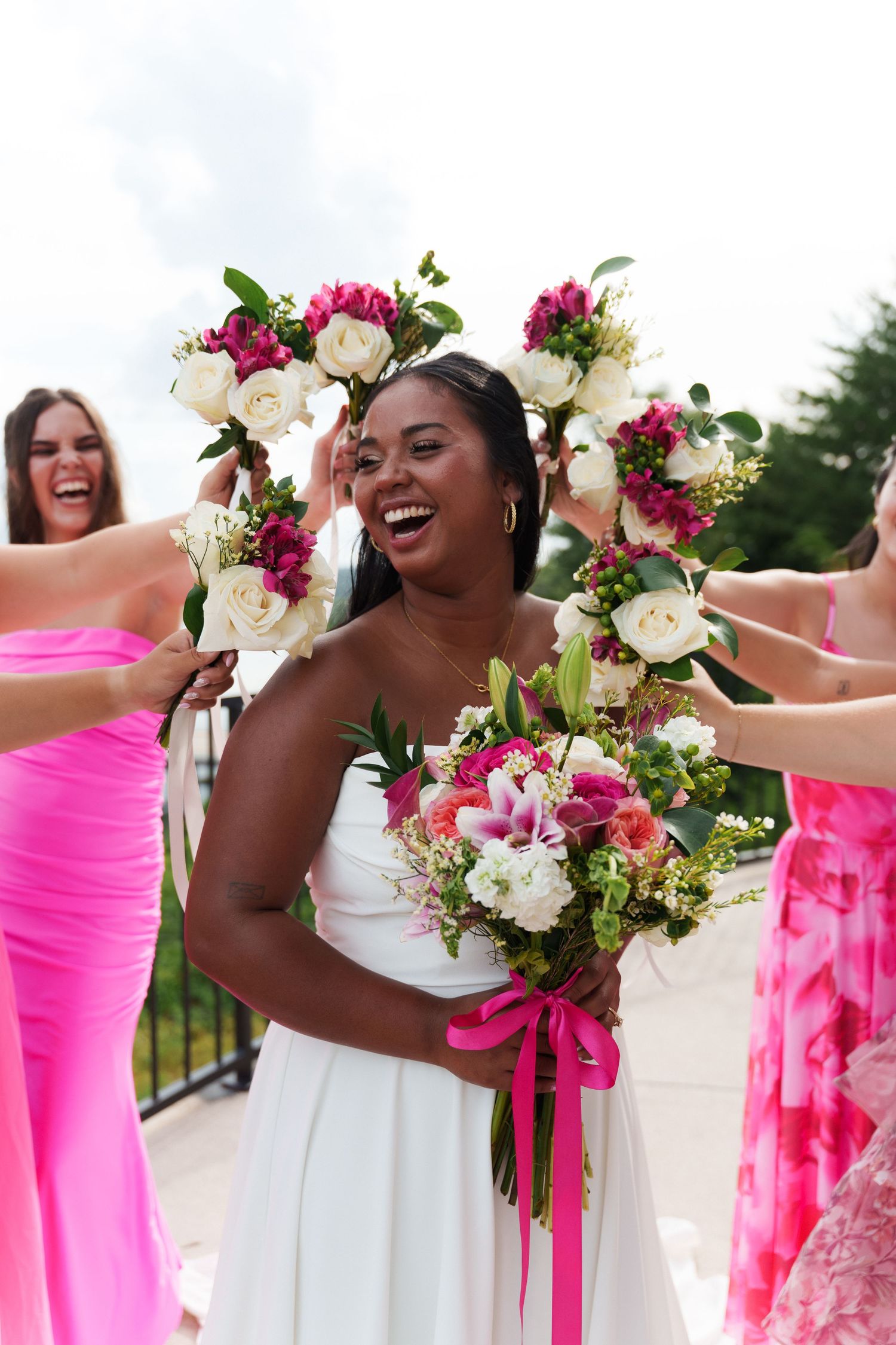 Joyful bride in white dress holds pink floral bouquet while bridesmaids in pink dresses surround her with flower arrangements.