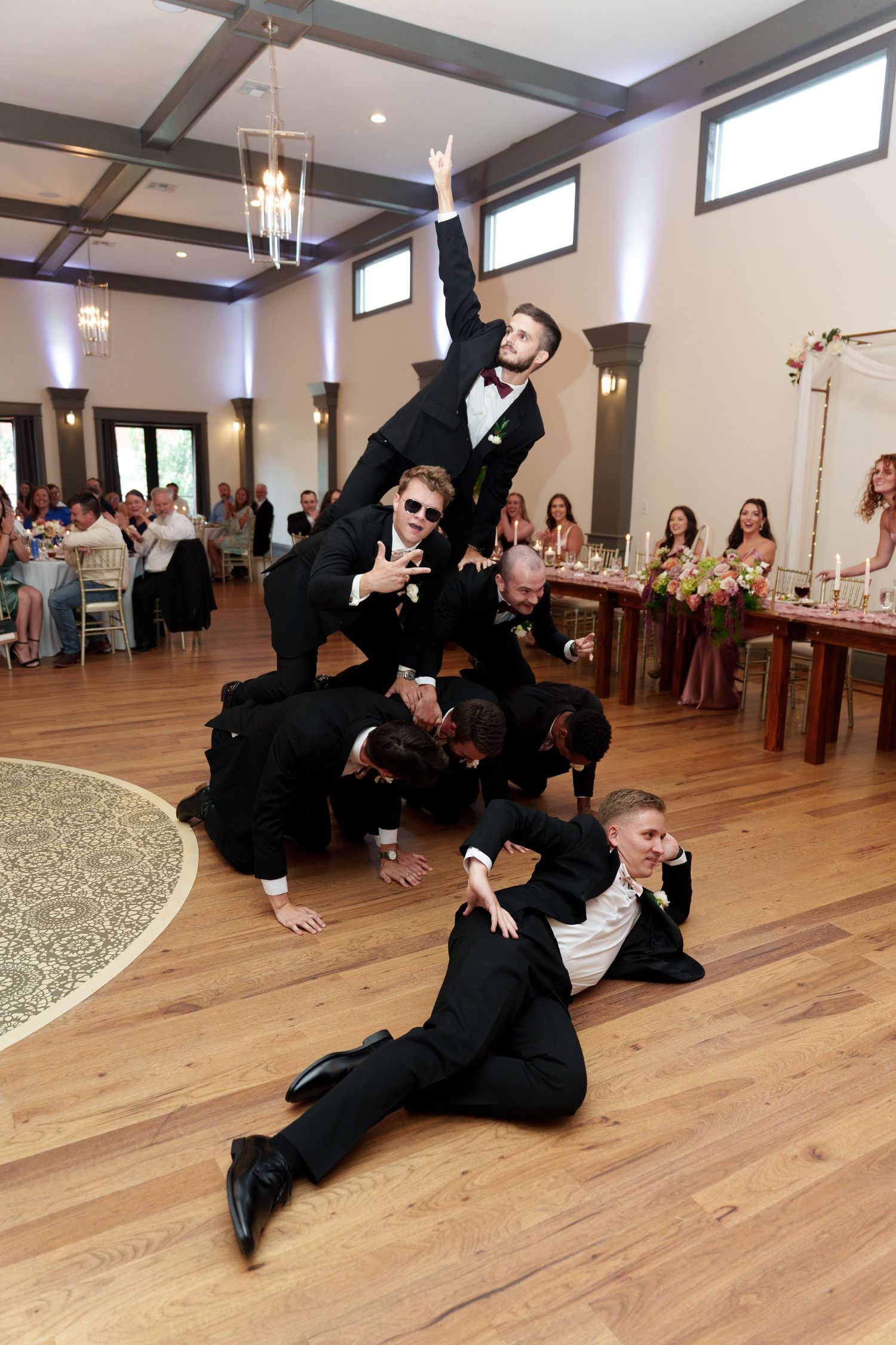 A wedding party performs a dynamic group dance move on a hardwood floor at the reception venue.