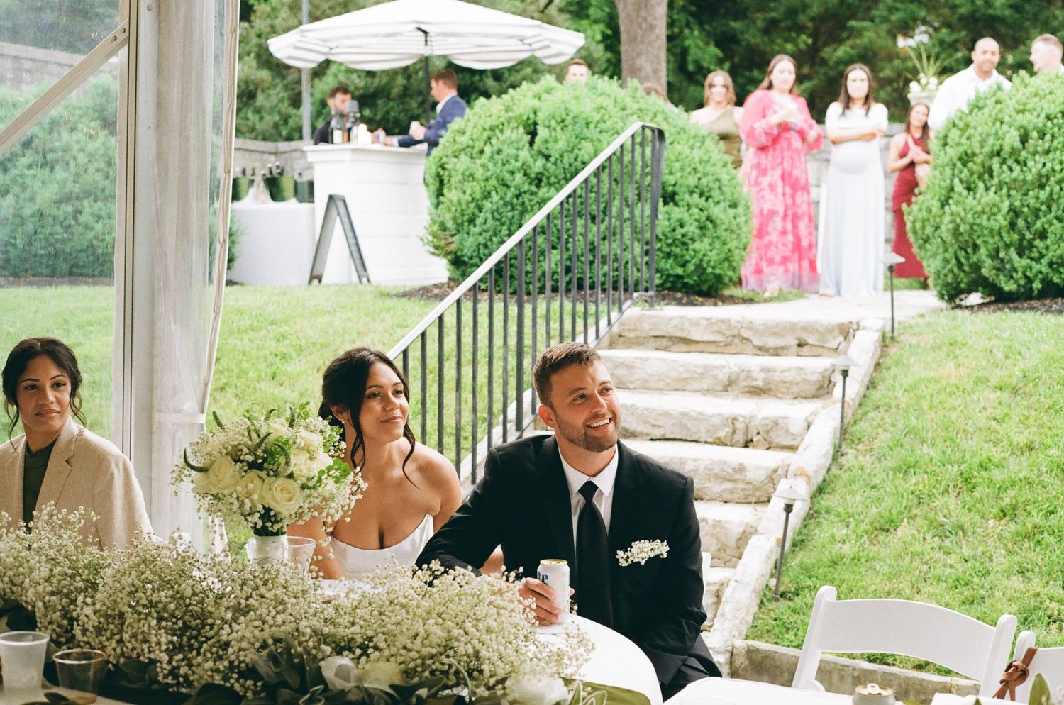 Bride and groom laughing at wedding reception table outdoors under tent with floral arrangements and guests in background.
