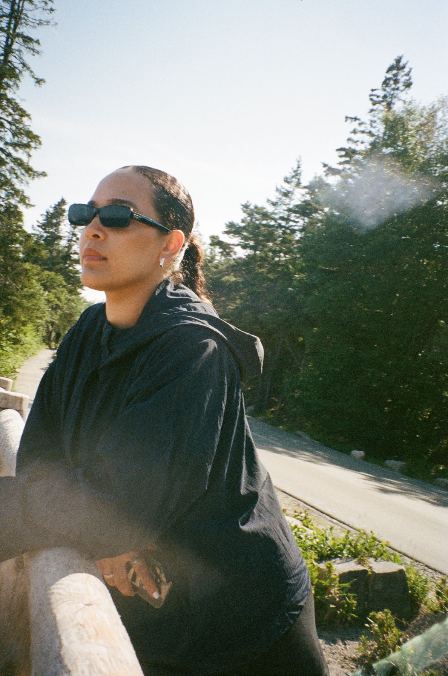 Woman in black hoodie and sunglasses leaning on wooden railing outdoors with trees and sunlight in background.