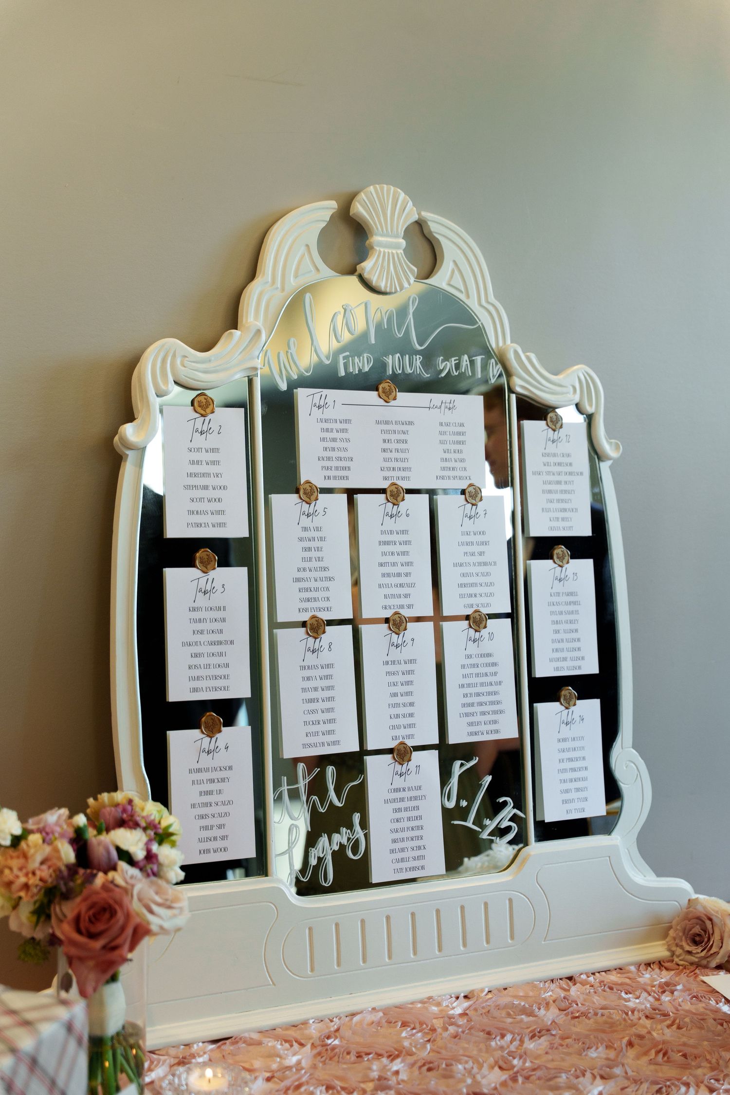 An ornate white mirror frame displays wedding seating arrangements with floral accents.