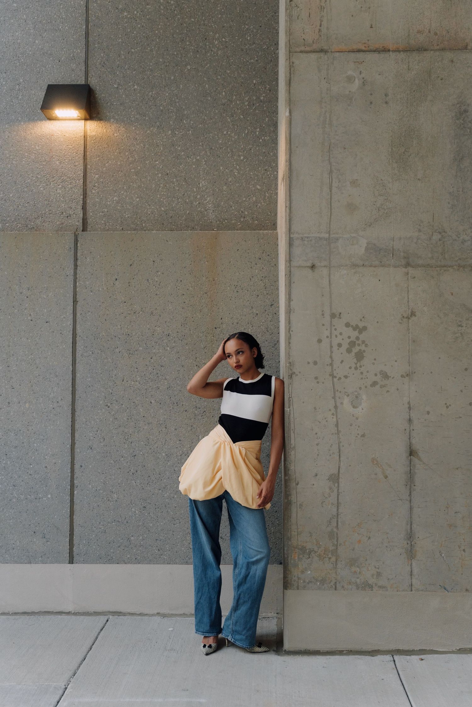 Woman in striped top, yellow draped skirt, and wide-leg jeans poses against concrete wall under wall light.