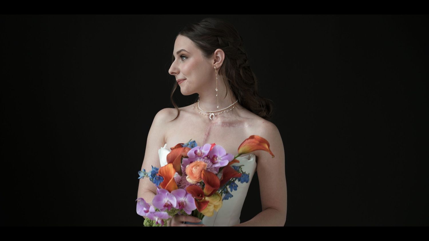 Elegant woman in strapless gown holding colorful floral bouquet, wearing statement necklace against dark background.