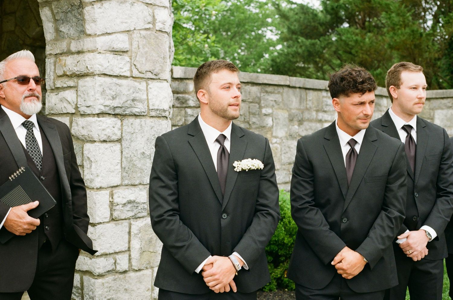Four groomsmen in black suits with dark ties stand outdoors at a wedding ceremony near a stone wall.