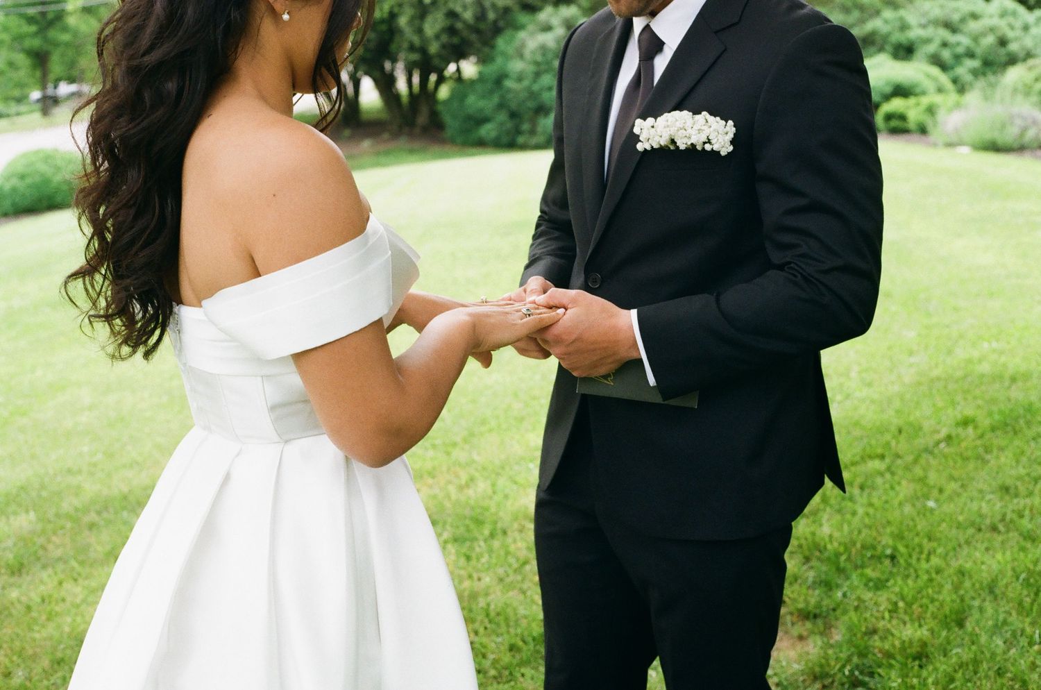 Bride in off-shoulder white gown exchanges rings with groom in black tuxedo during outdoor wedding ceremony.