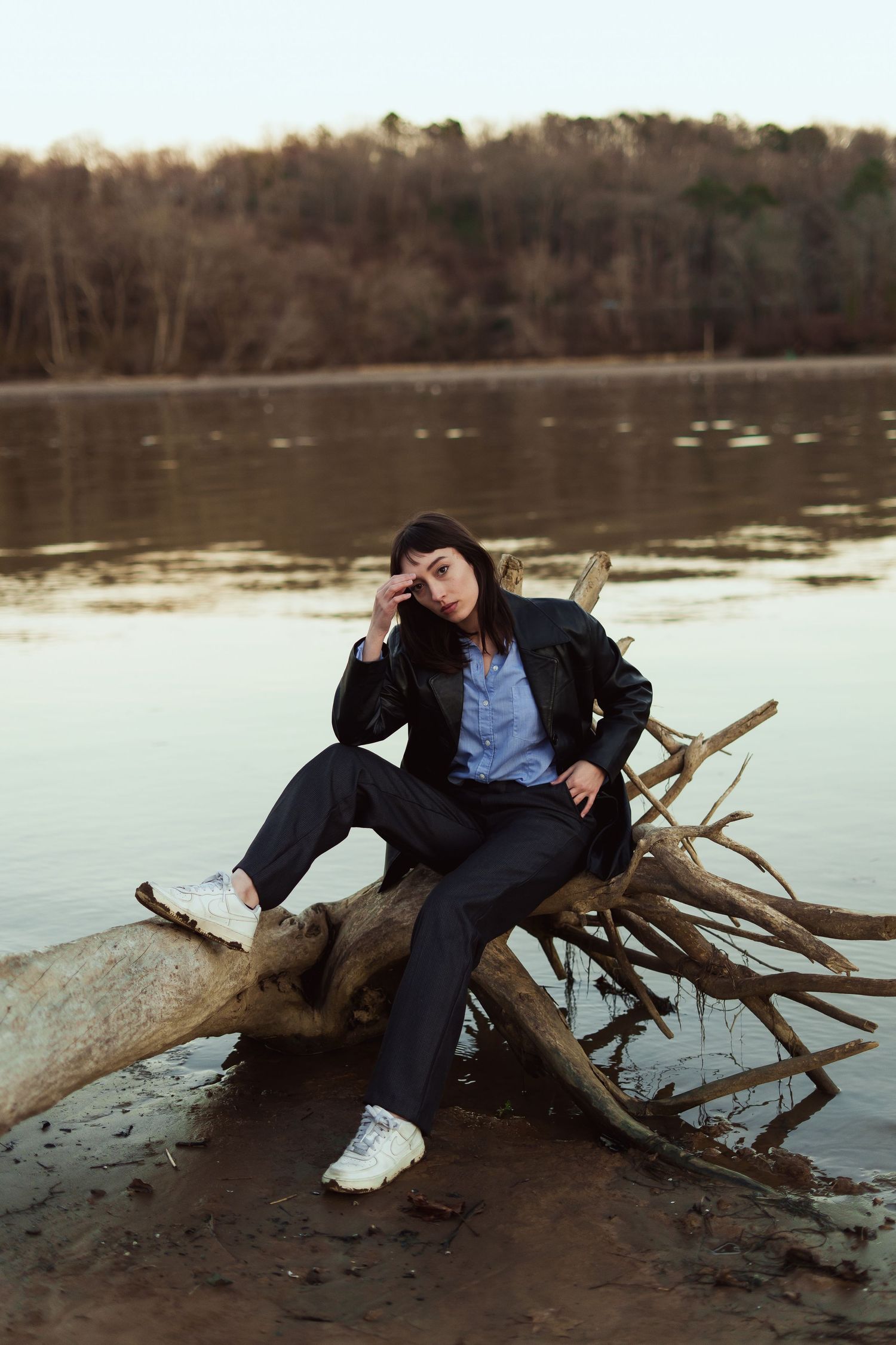 Person relaxing on fallen tree trunk by tranquil lake during sunset wearing black jacket and jeans with white sneakers.