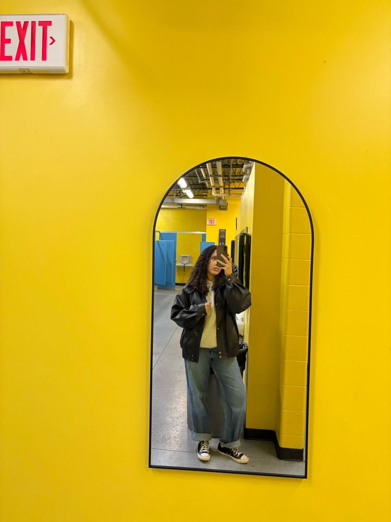 Person taking mirror selfie in black leather jacket and wide-leg jeans against bright yellow wall near exit sign.