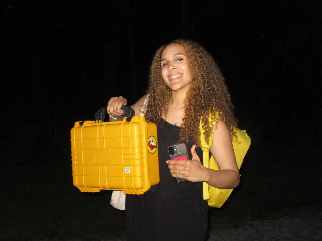Young woman with curly hair smiling outdoors at night, holding a yellow Pelican case and wearing a yellow backpack.