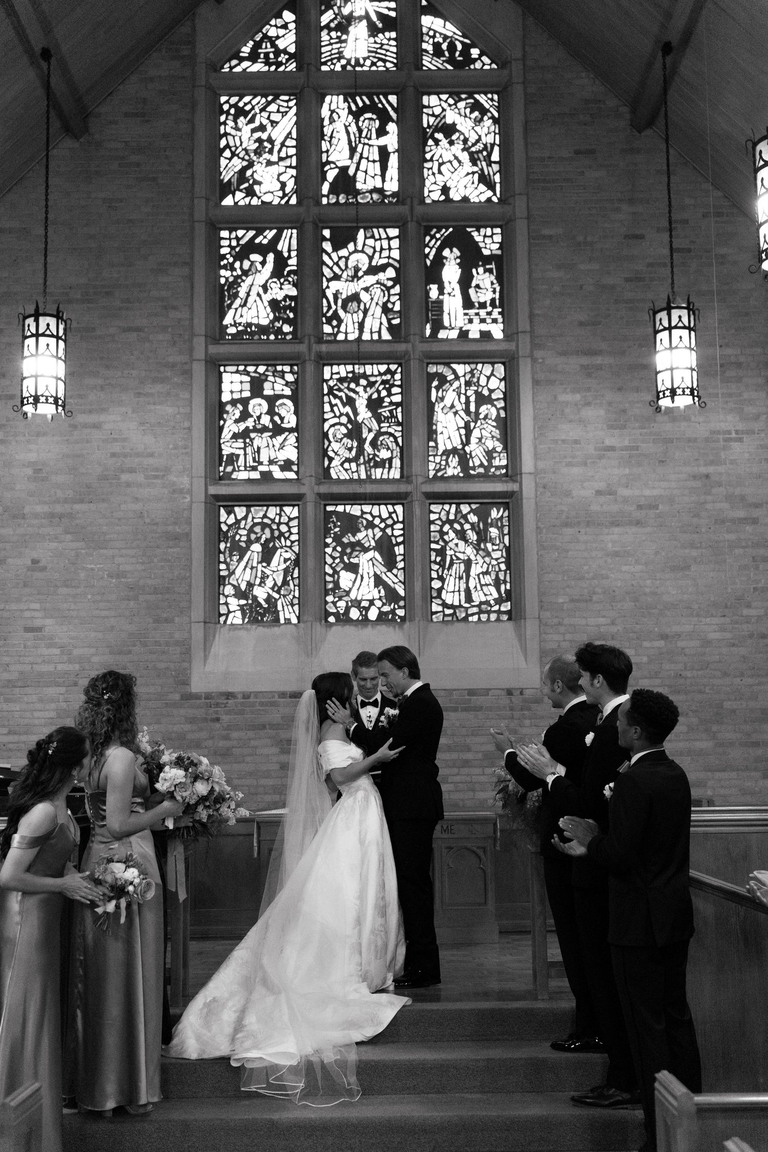 A black and white wedding scene showing four couples dipping and kissing simultaneously in an elegant church setting.
