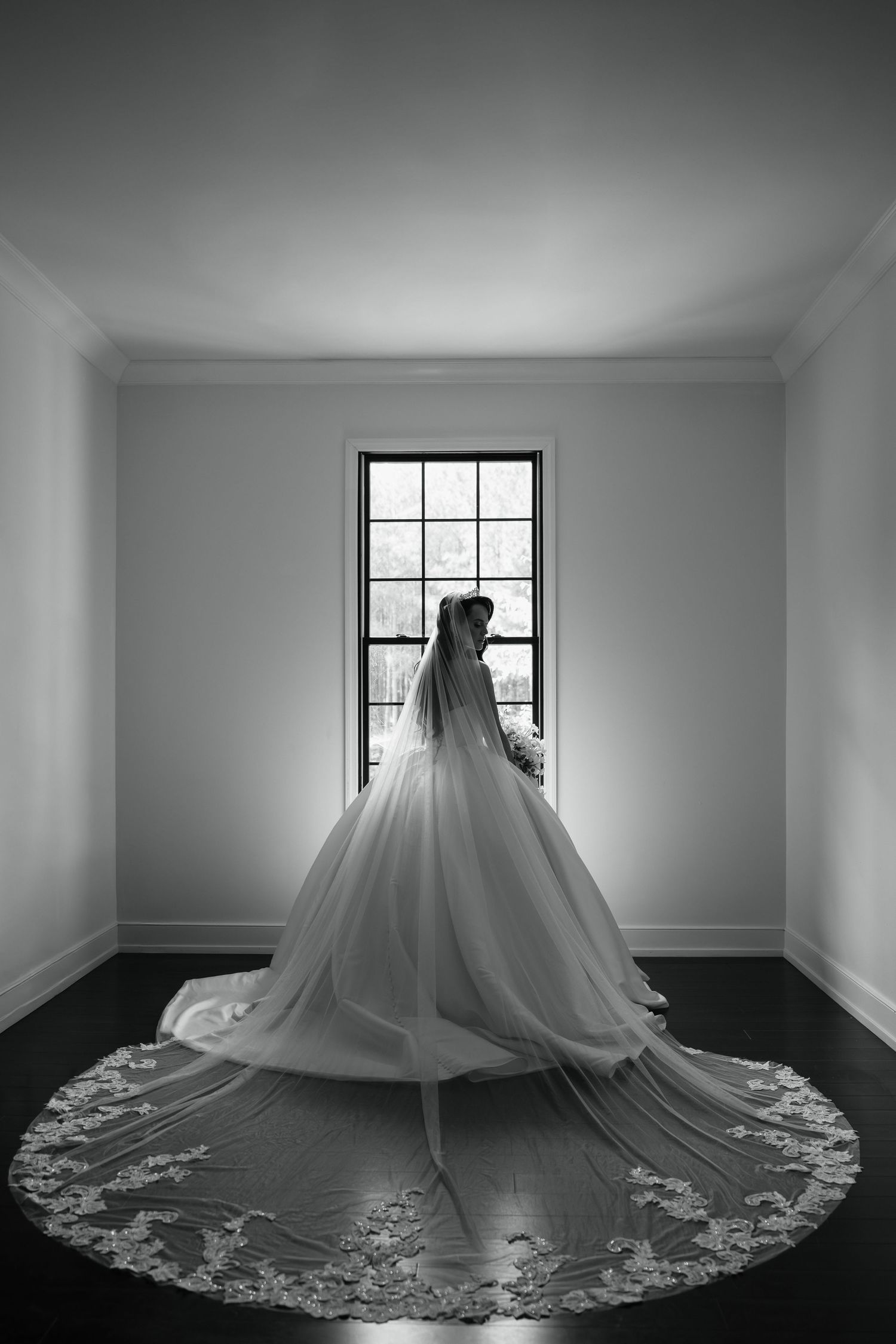 Black and white photo of a bride in a flowing ballgown with lace-trimmed cathedral veil, standing before a tall window.